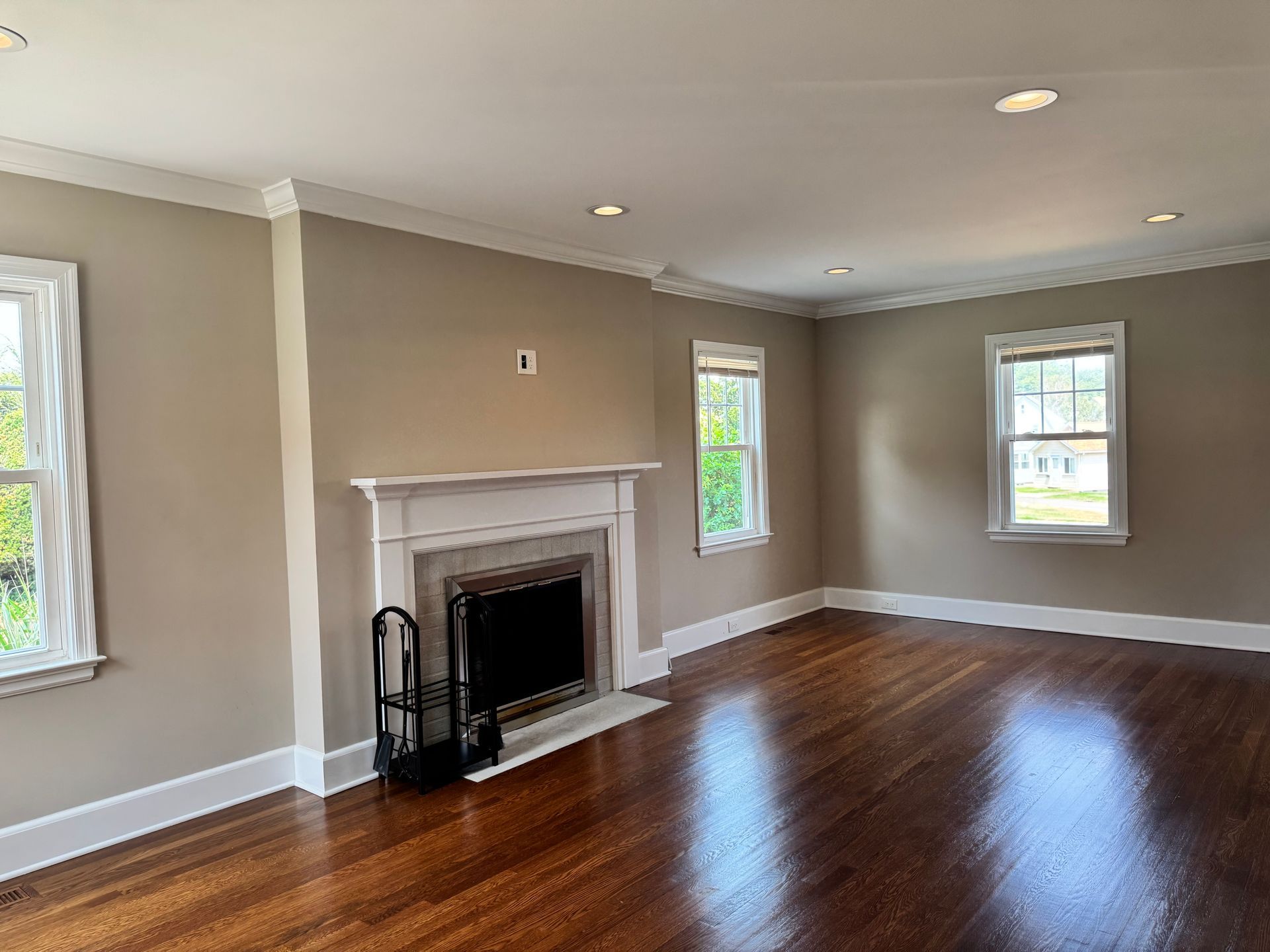 Empty living room with fireplace, hardwood floors, and windows. Beige walls, white trim.