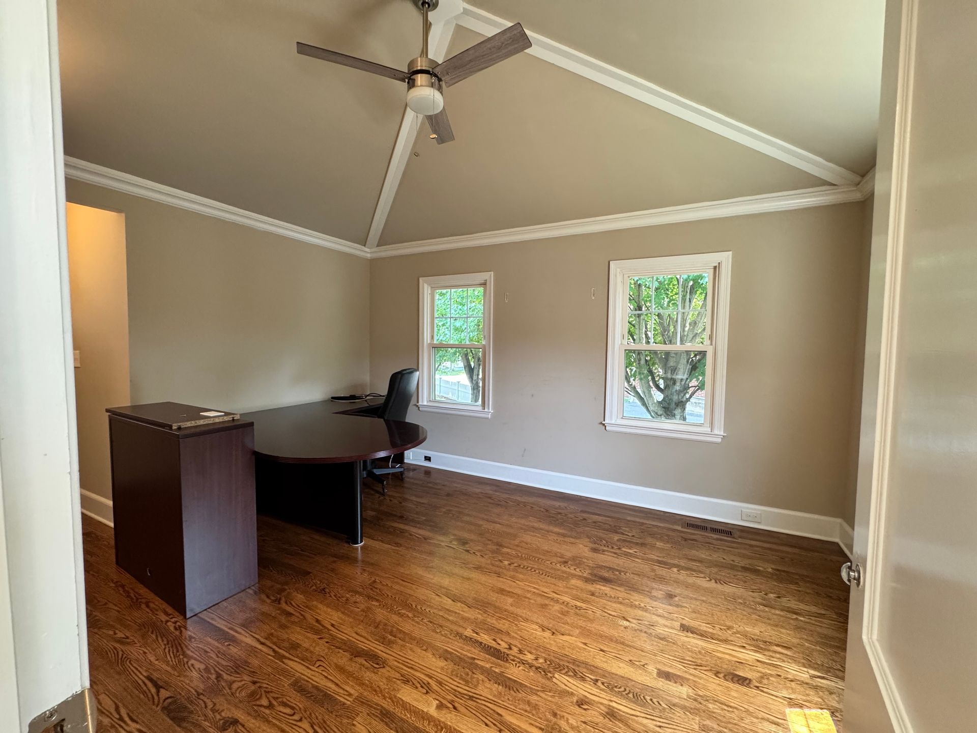 Empty office with dark wood desk, two windows, vaulted ceiling, and wood floors.