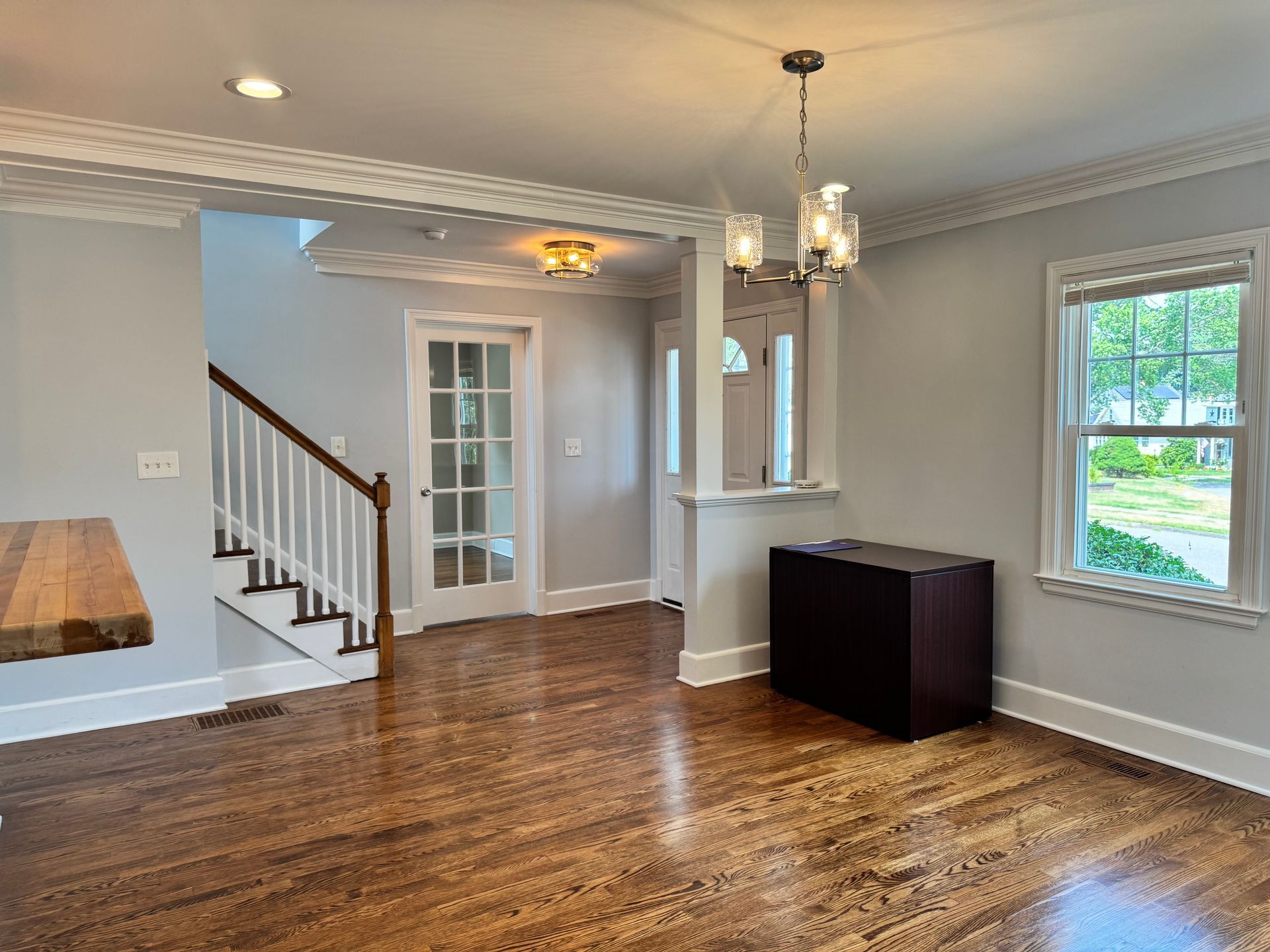 Interior of a house with hardwood floors, light gray walls, a staircase, and a dining area with a chandelier.