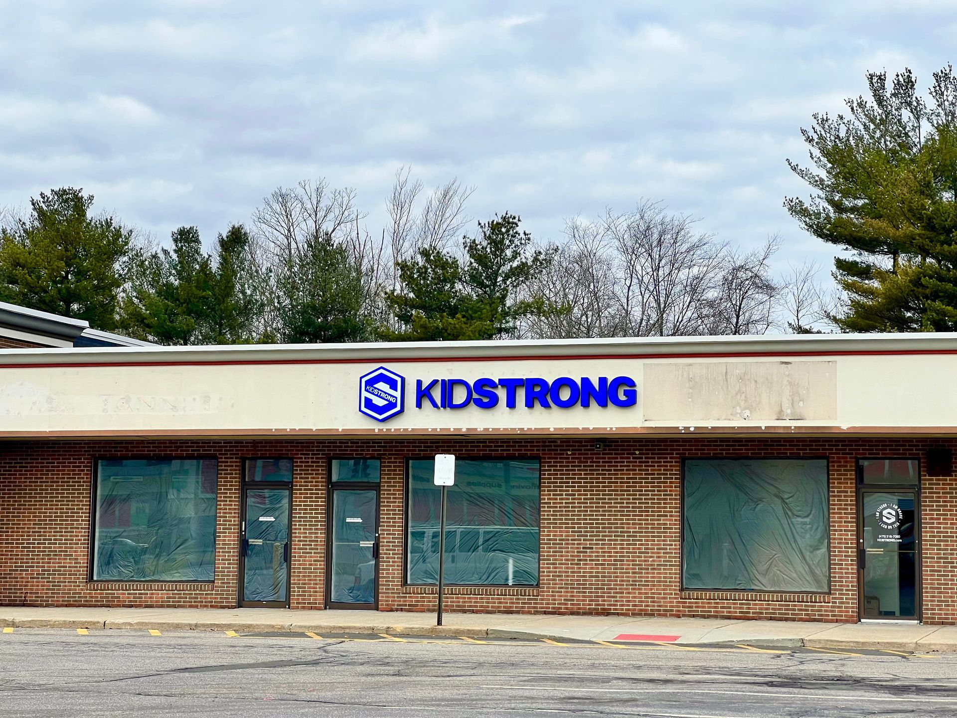 Exterior view of a KidStrong storefront in a brick building with a blue sign. Windows are boarded up.