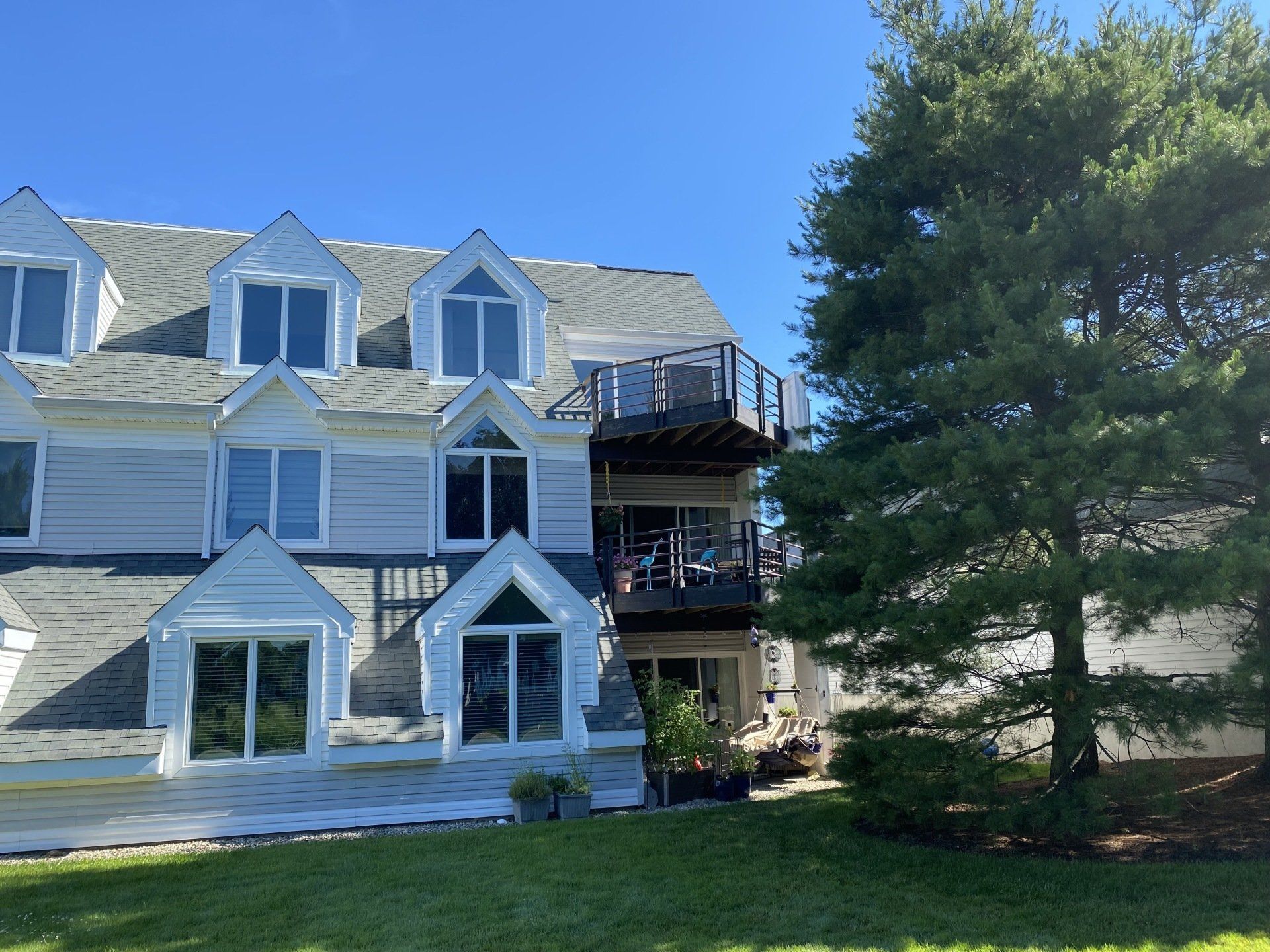 Light blue apartment building with multiple gabled dormer windows and a wooden deck, next to a large evergreen tree