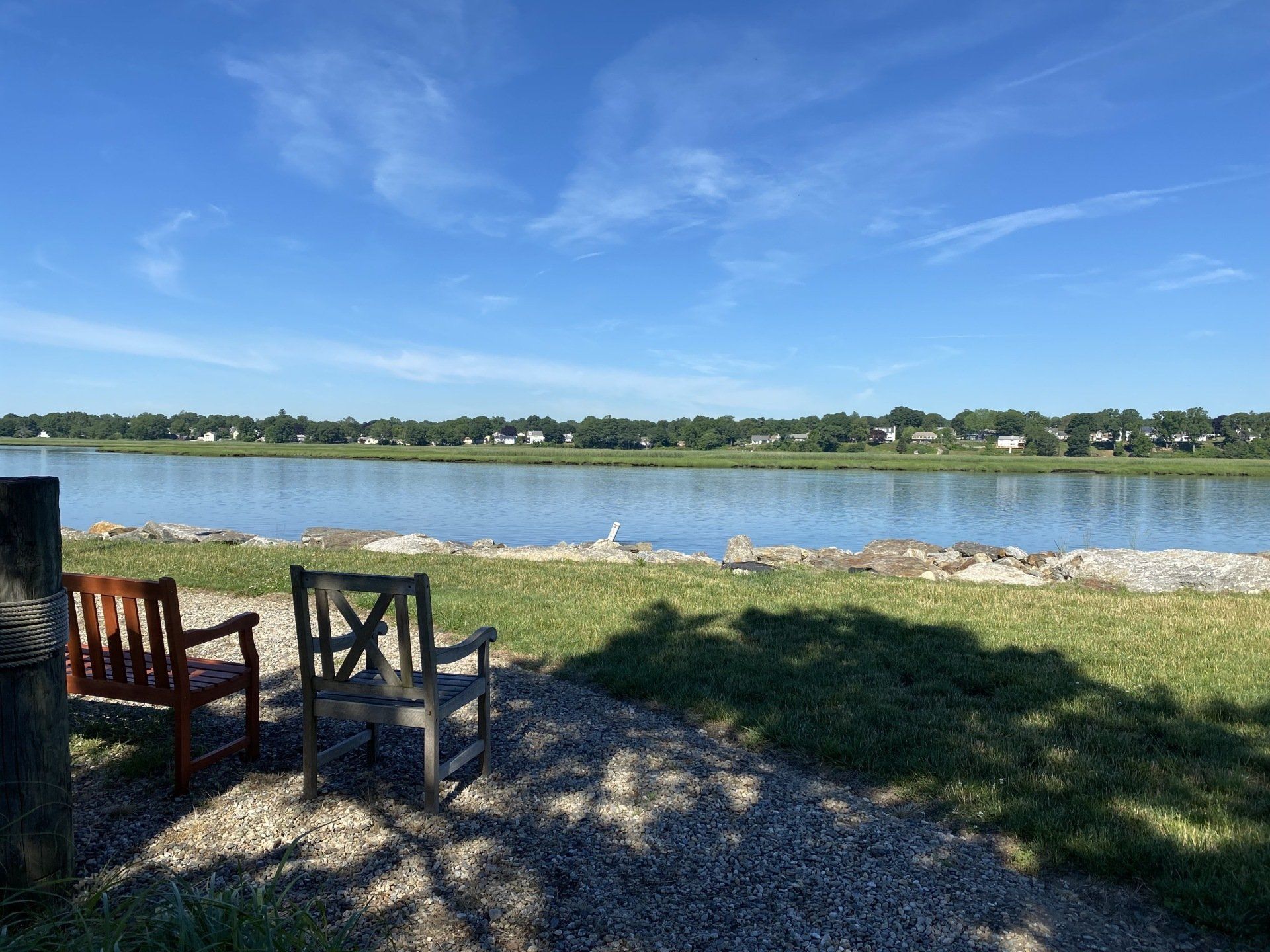 Two wooden chairs sit near a waterfront with a blue sky and distant buildings.
