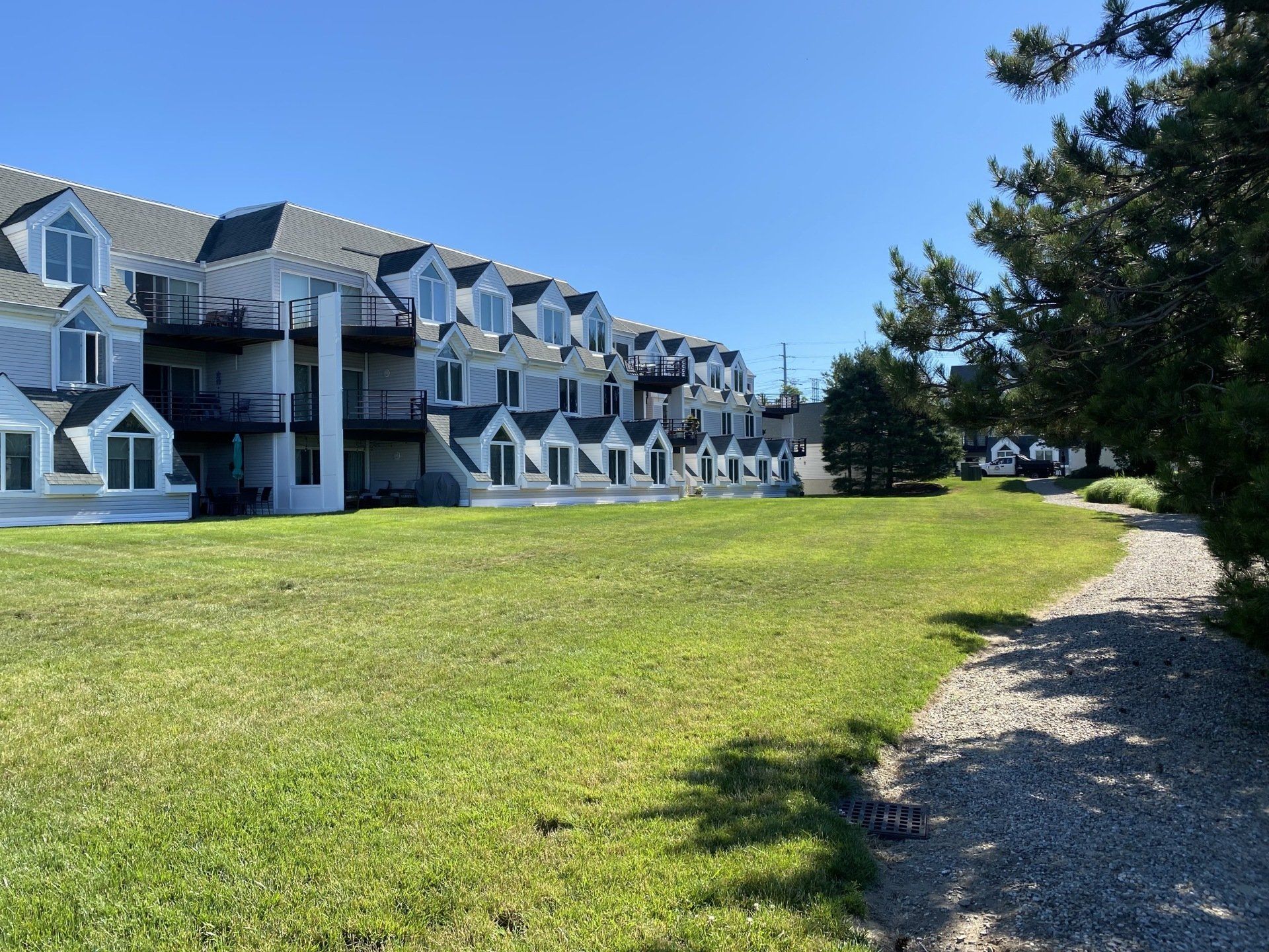 Apartment building with dormer windows, green lawn, and gravel path on a sunny day.