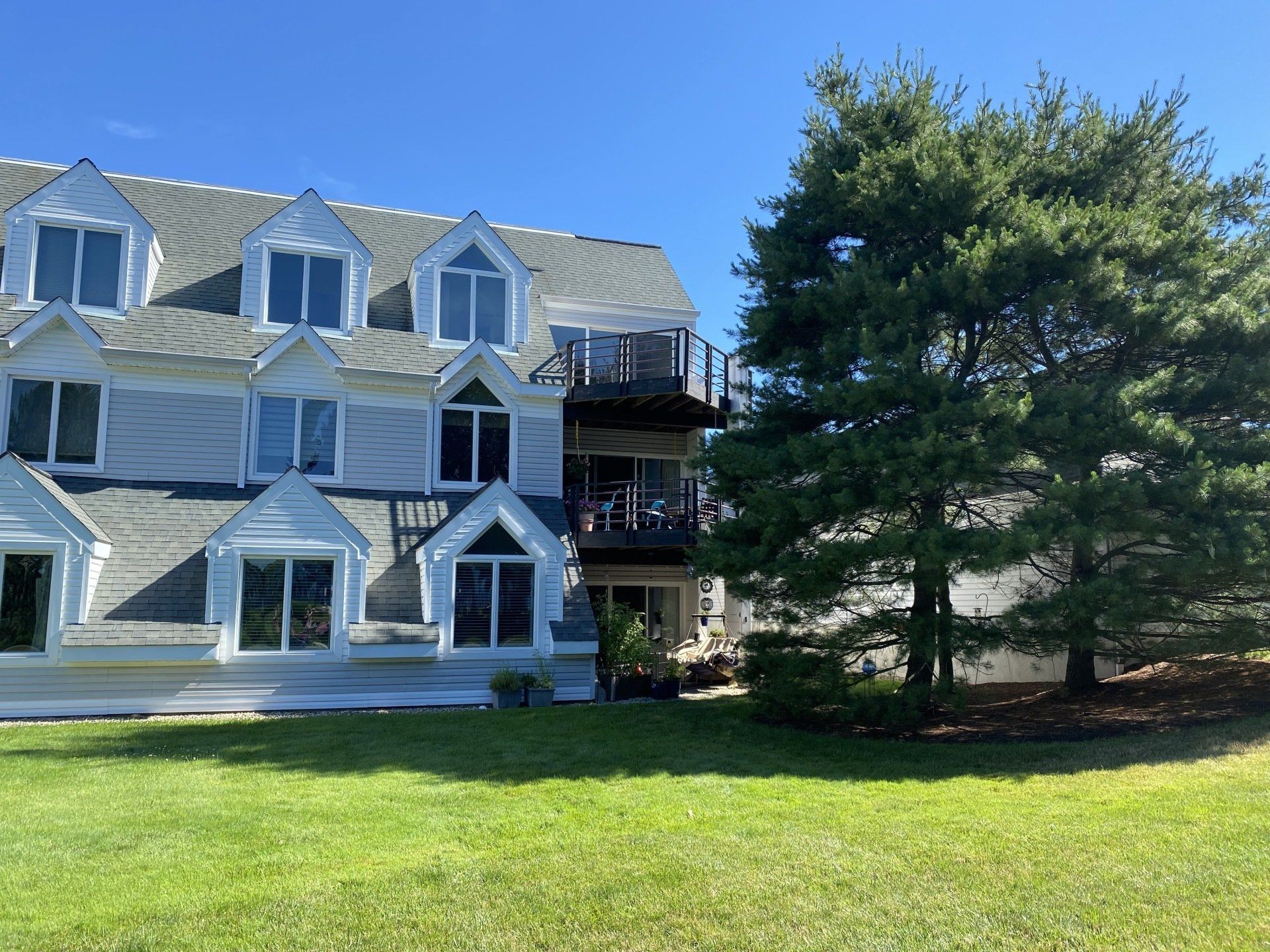 Light blue multi-story building with multiple windows, a black deck, and a tall green tree on a grassy lawn.