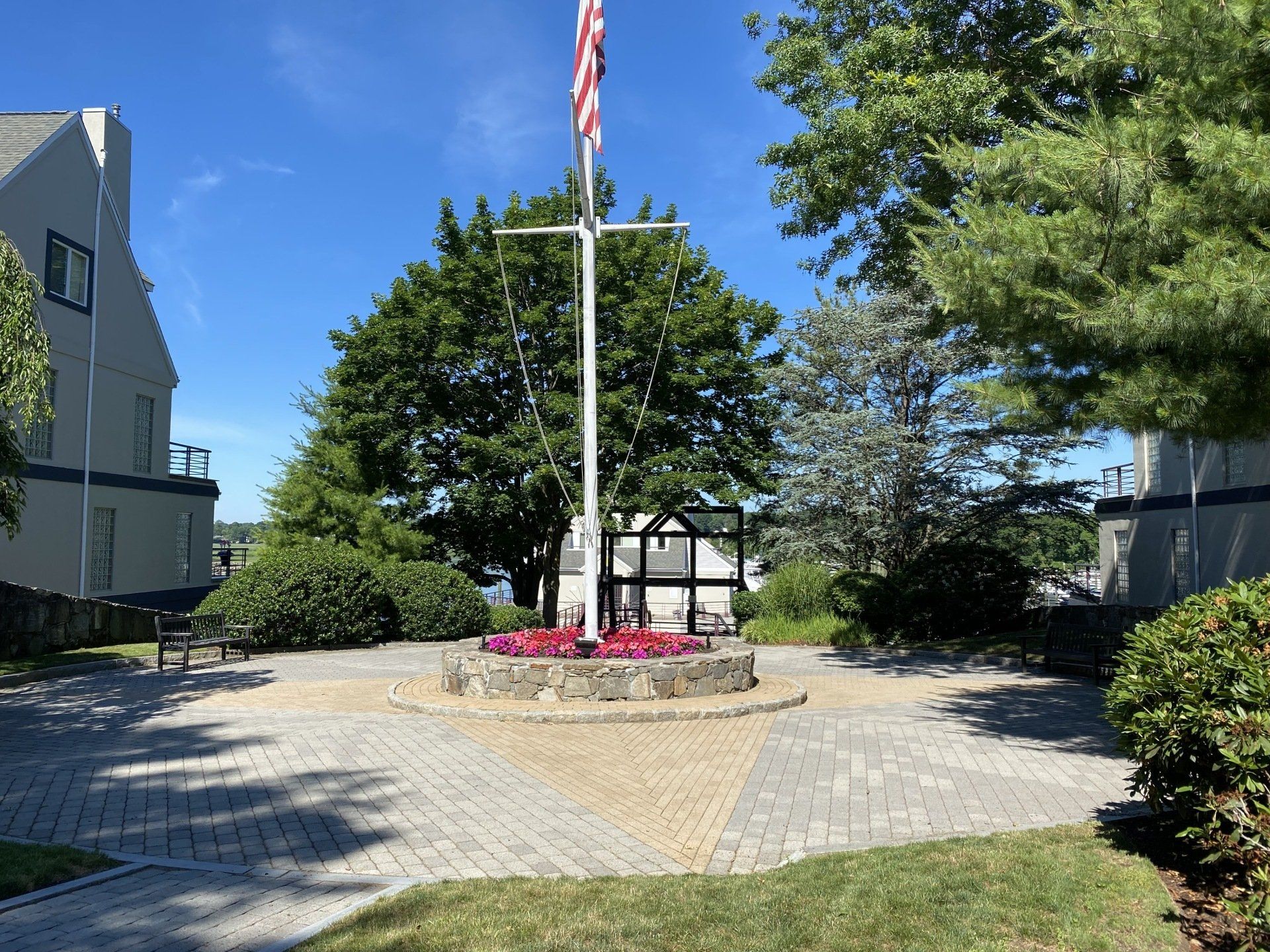Flagpole in a circular brick plaza with flower bed, flanked by buildings and trees on a sunny day.