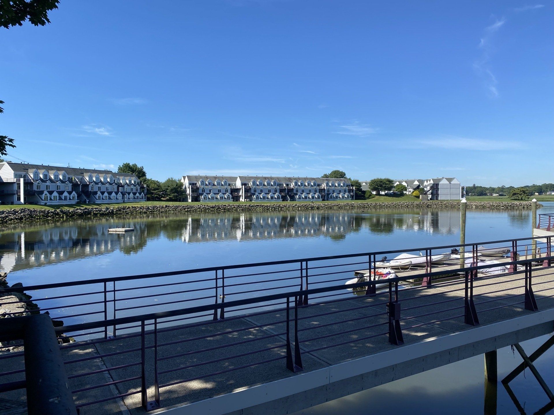 Waterfront view: row of buildings reflected in calm water, blue sky, dock in foreground.