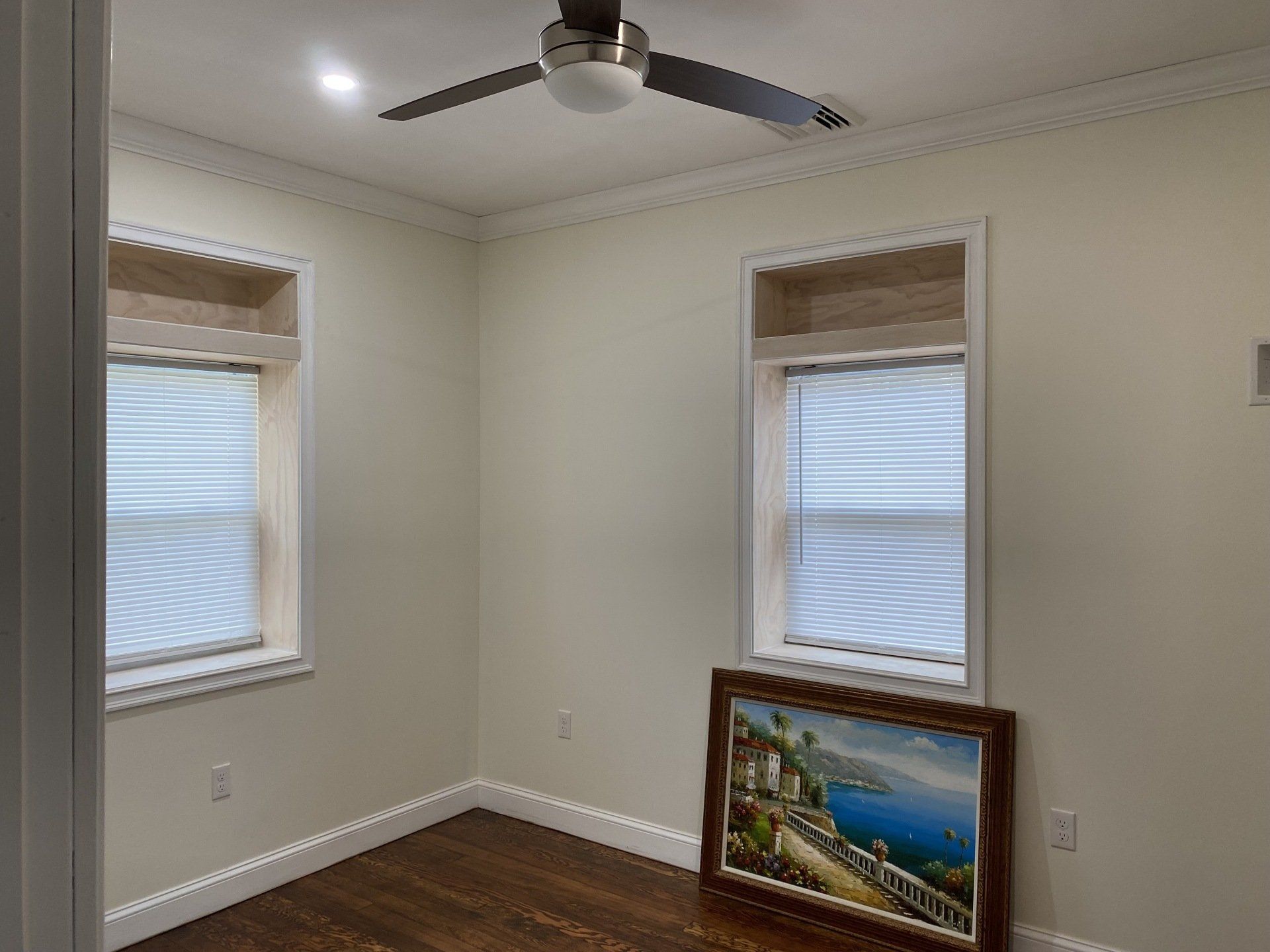 Empty room with two windows, a ceiling fan, and a painting leaning against the wall. Wooden floor, cream-colored walls.