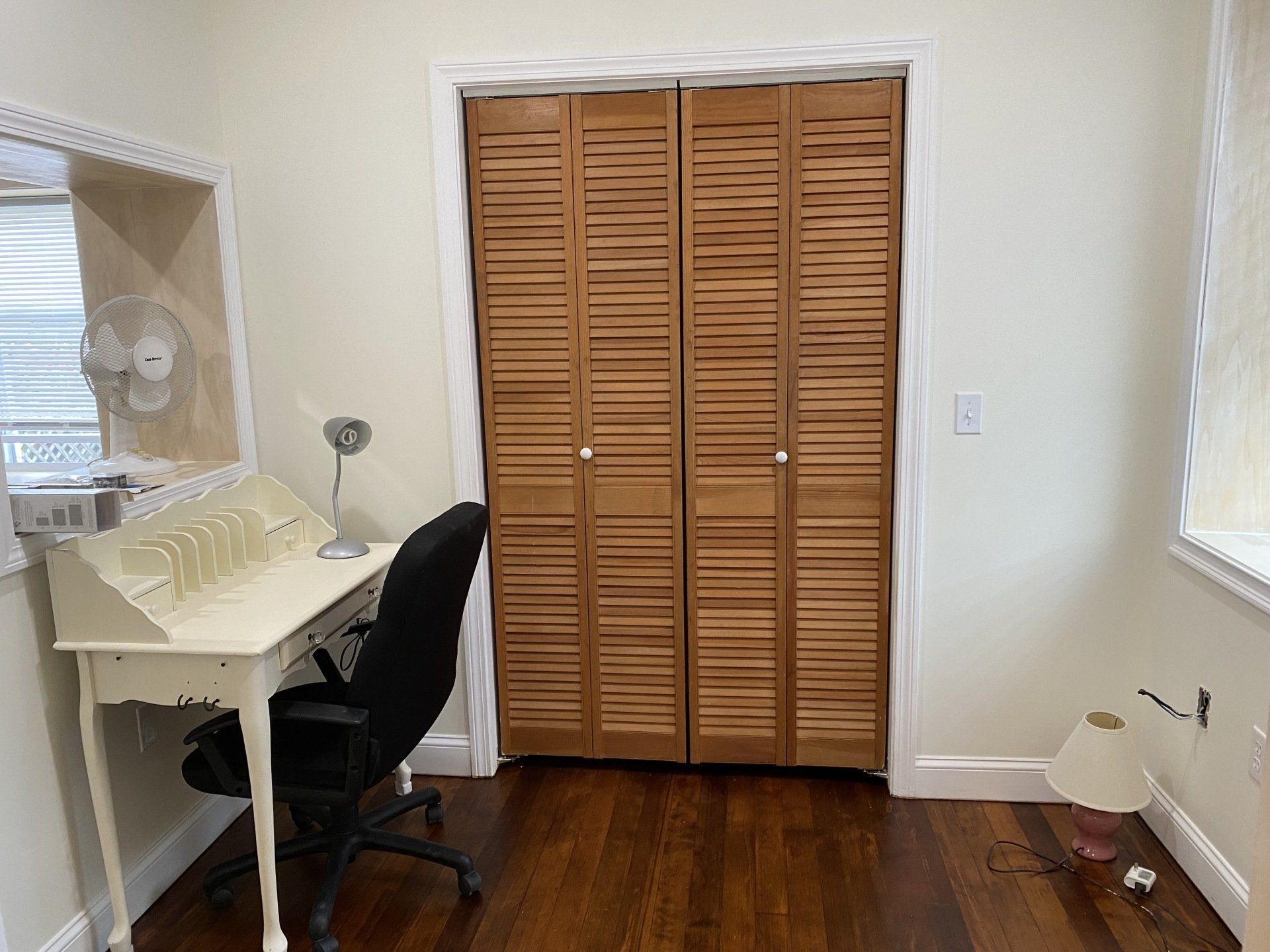 A small home office with a desk, closet, and chair. Dark wood flooring and off-white walls.