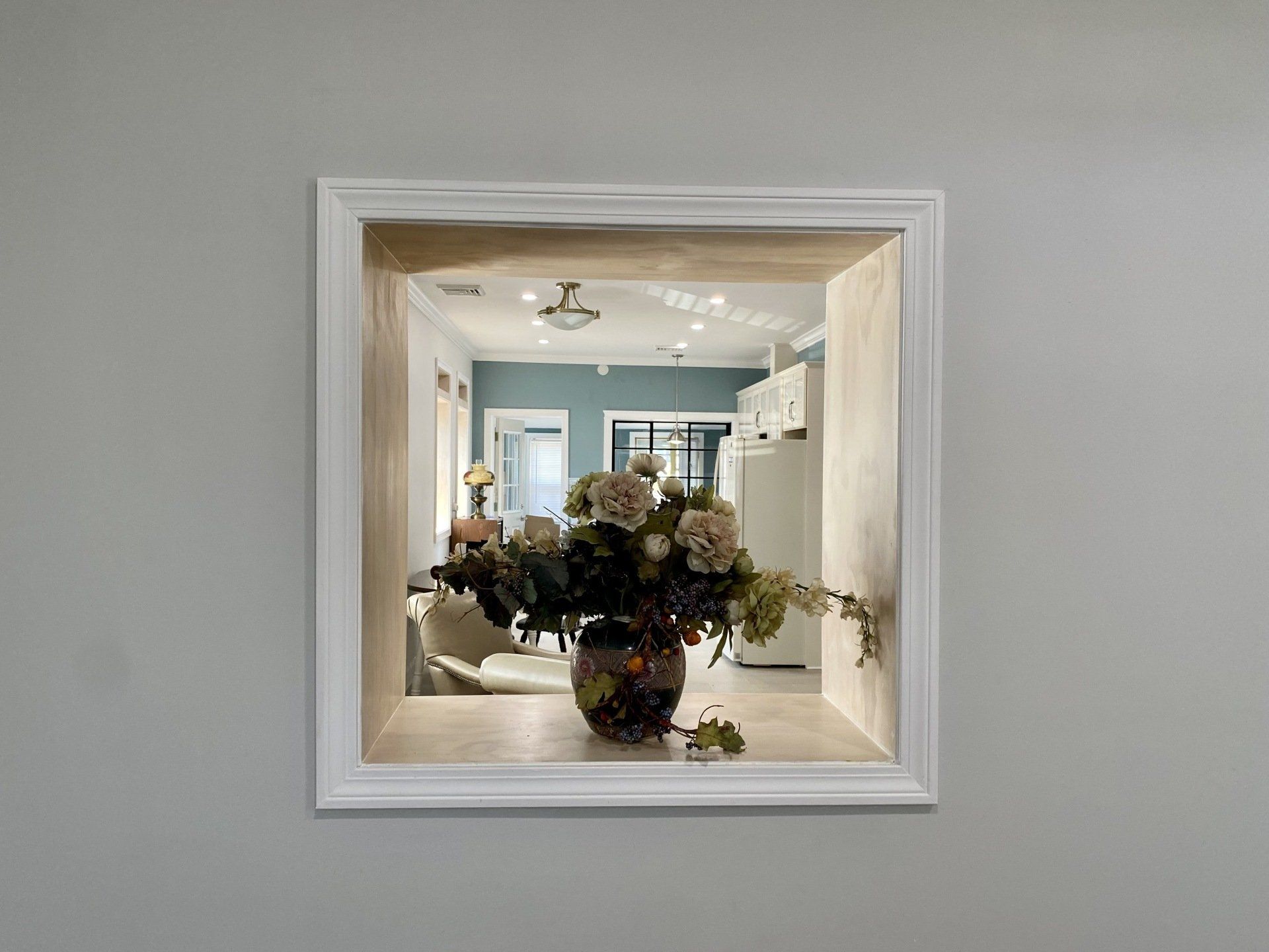 Square mirror on a gray wall; a vase of flowers sits on a shelf reflecting a room.