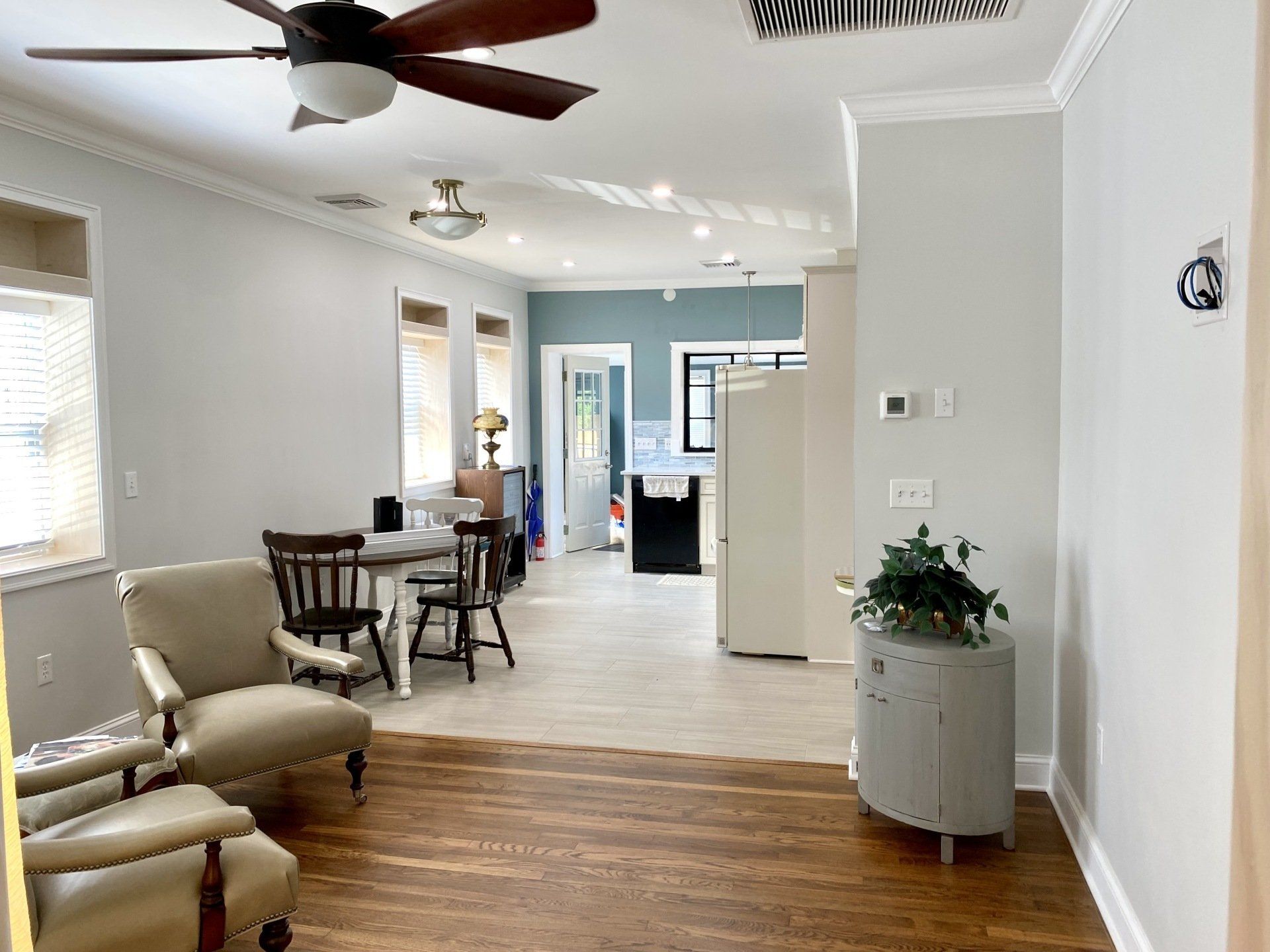 Living room with hardwood floors, chairs, and table. Kitchen with white appliances. Pale blue walls.