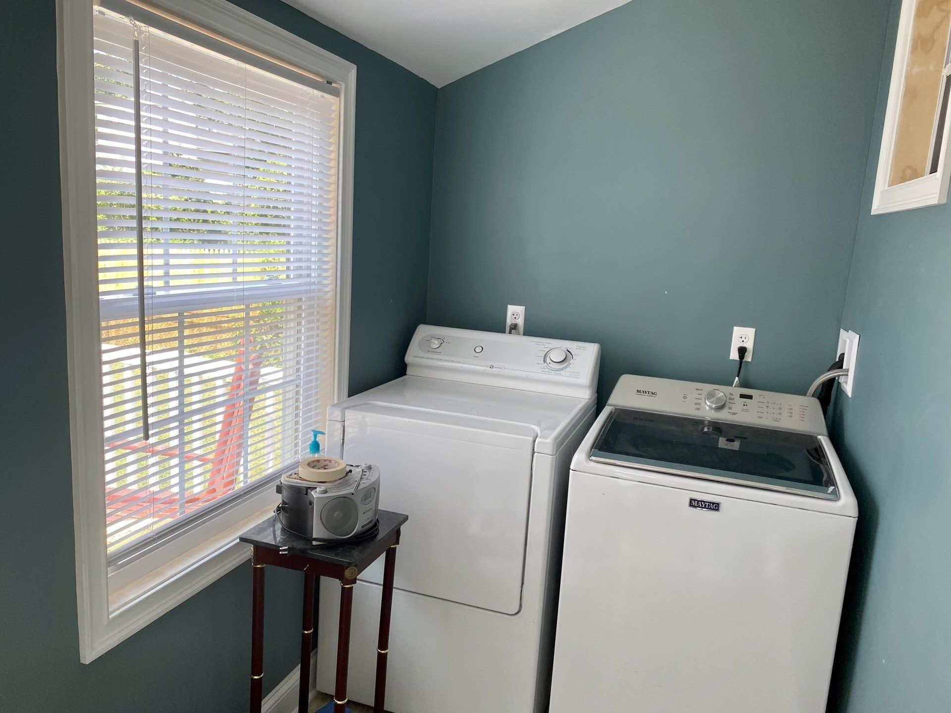 Laundry room with white washer and dryer, blue walls, a window with blinds, and a small table.