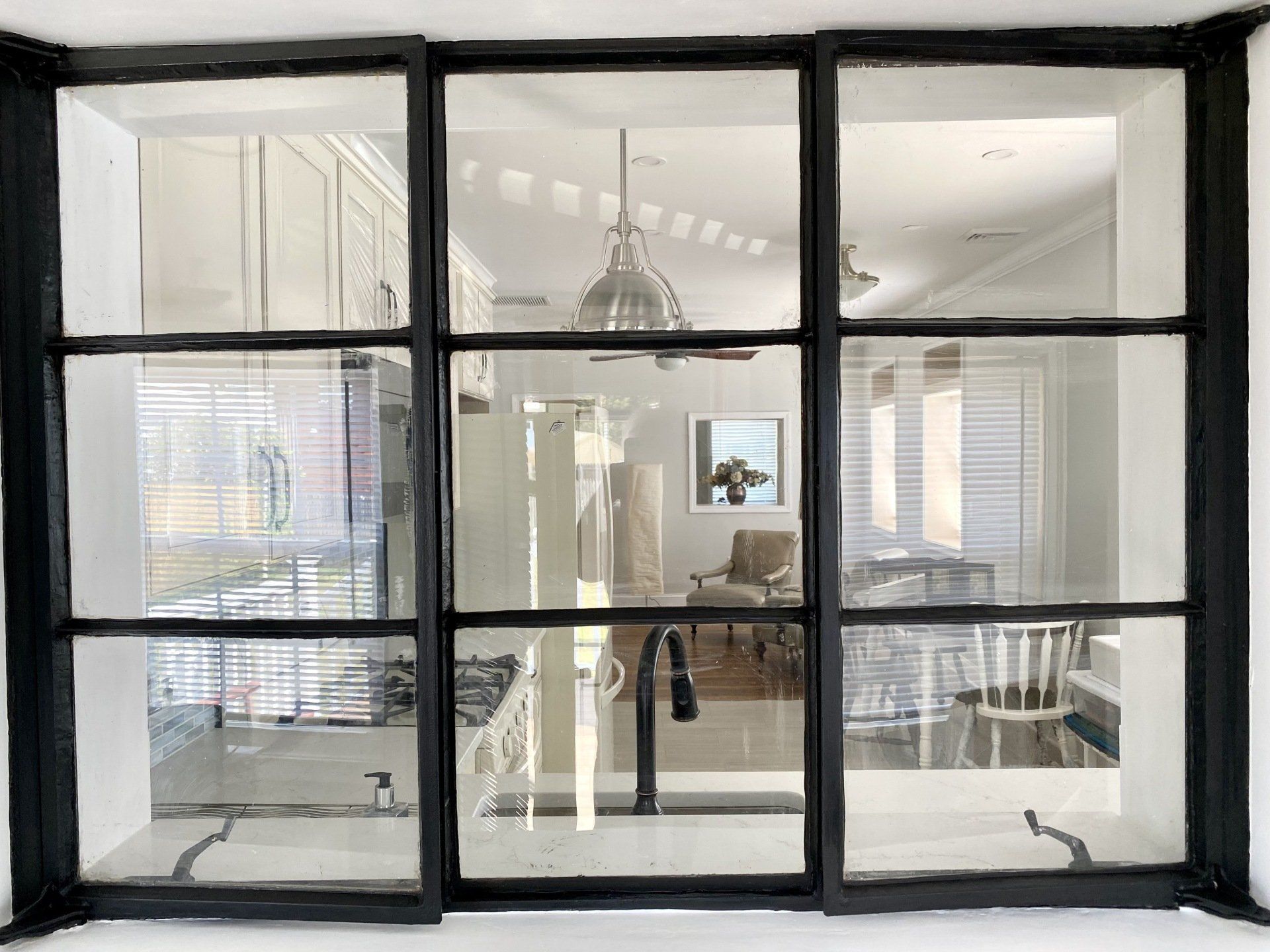 Black-framed window looking into a bright kitchen; black and white cabinets, large metal pendant lights