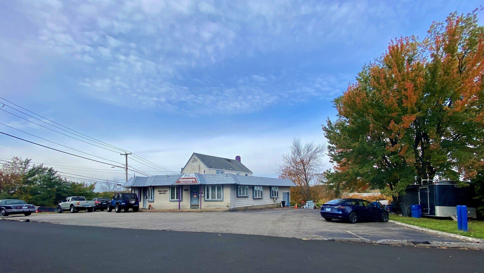 A low, light-colored building with a gravel parking lot and a blue sky in the background; a few vehicles are parked in front.