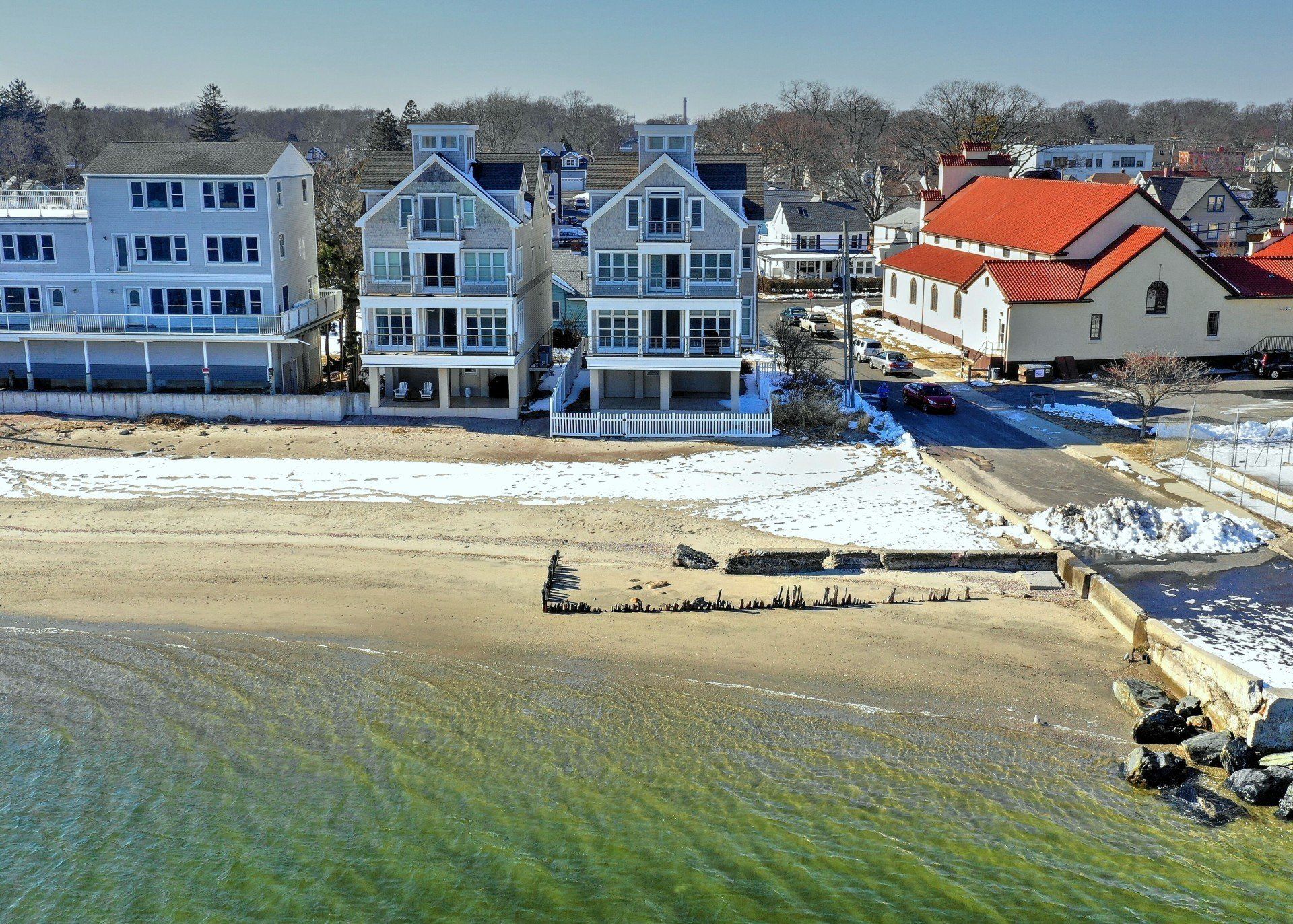 Beachfront homes, gray and white, near a church with a red roof, winter scene with snow.