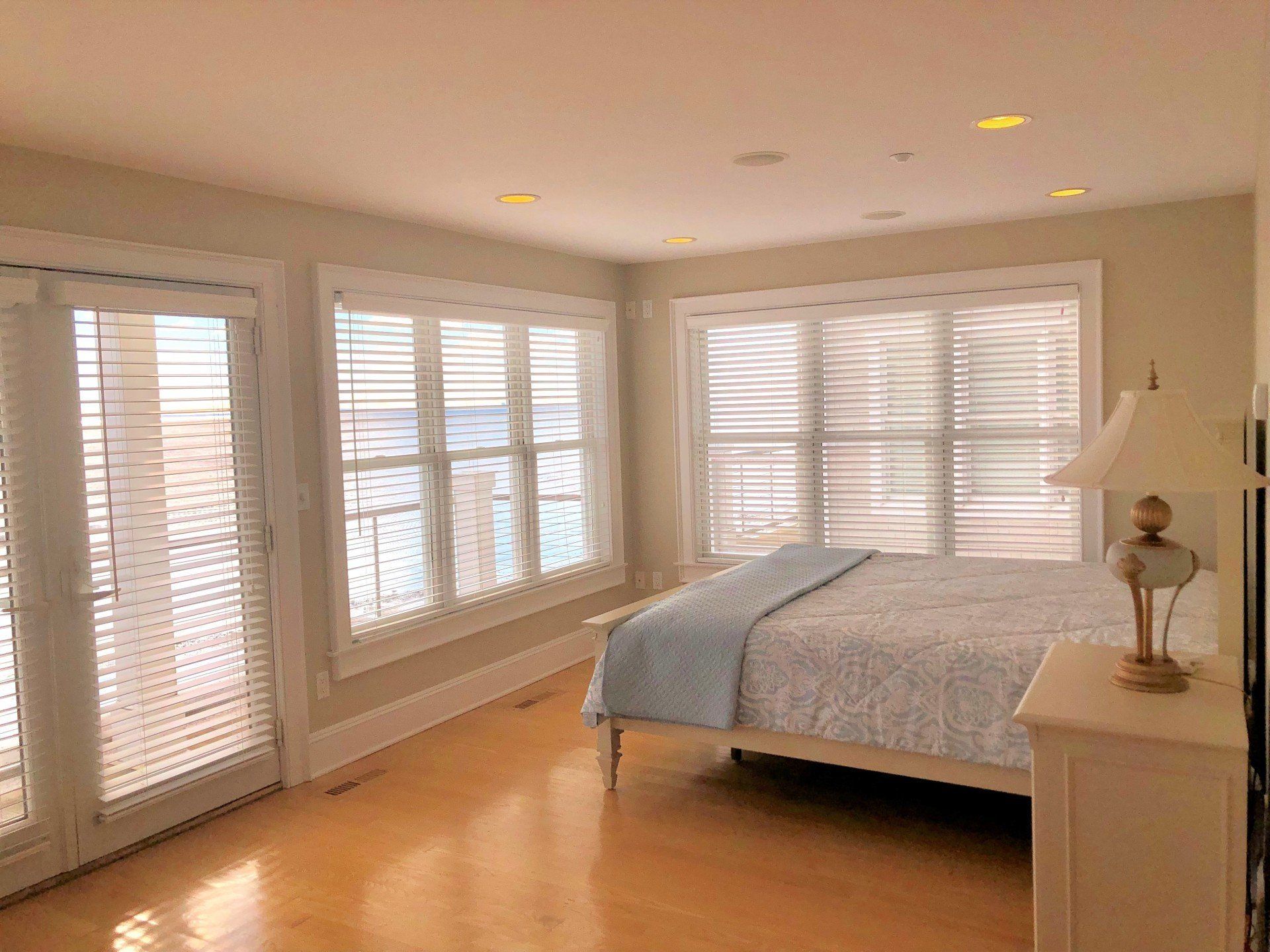 Bright bedroom with light wood floors, bed with blue blanket, large windows with blinds.