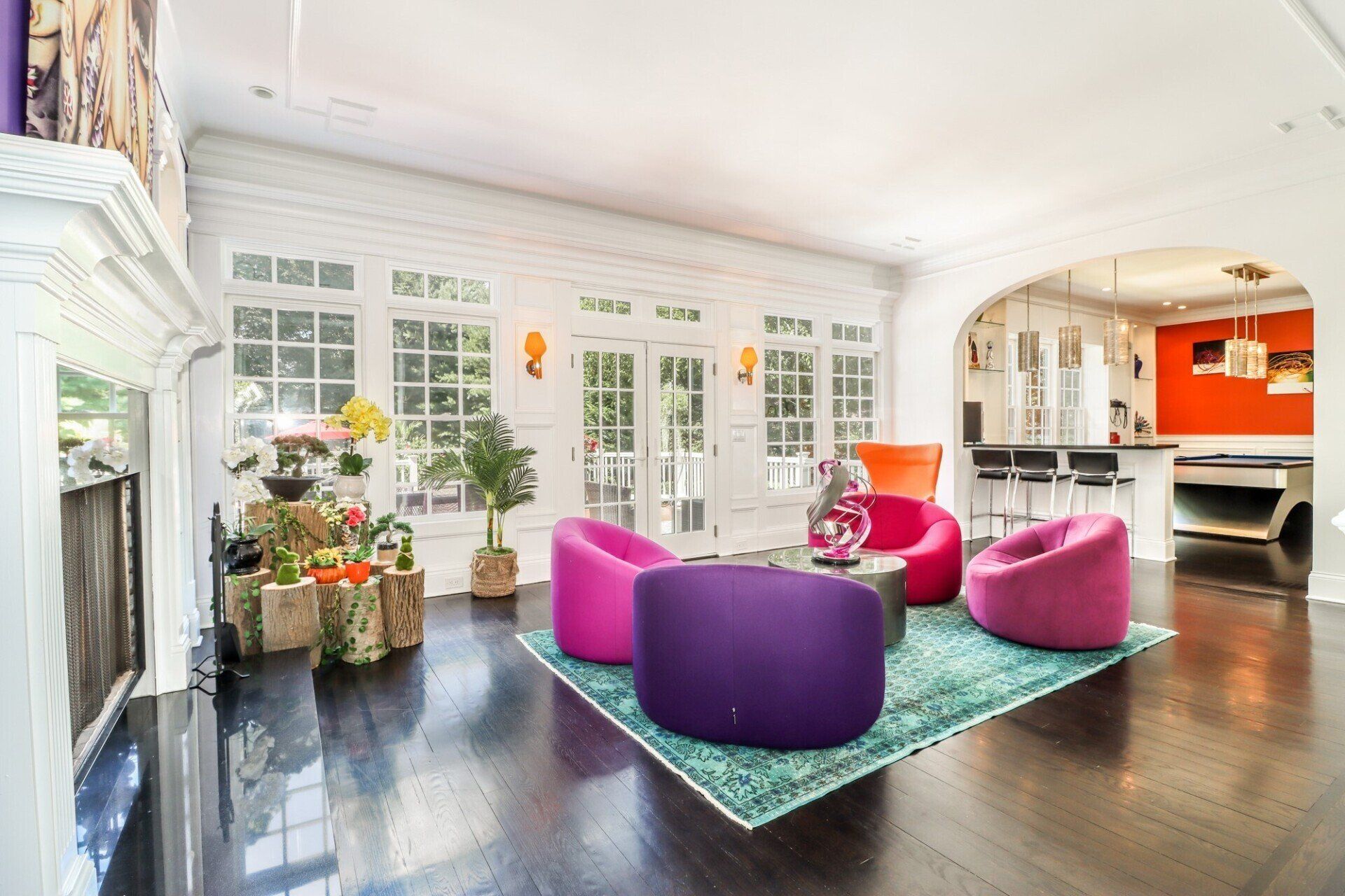 Bright, modern living room with colorful chairs on a turquoise rug, hardwood floors, and large windows.