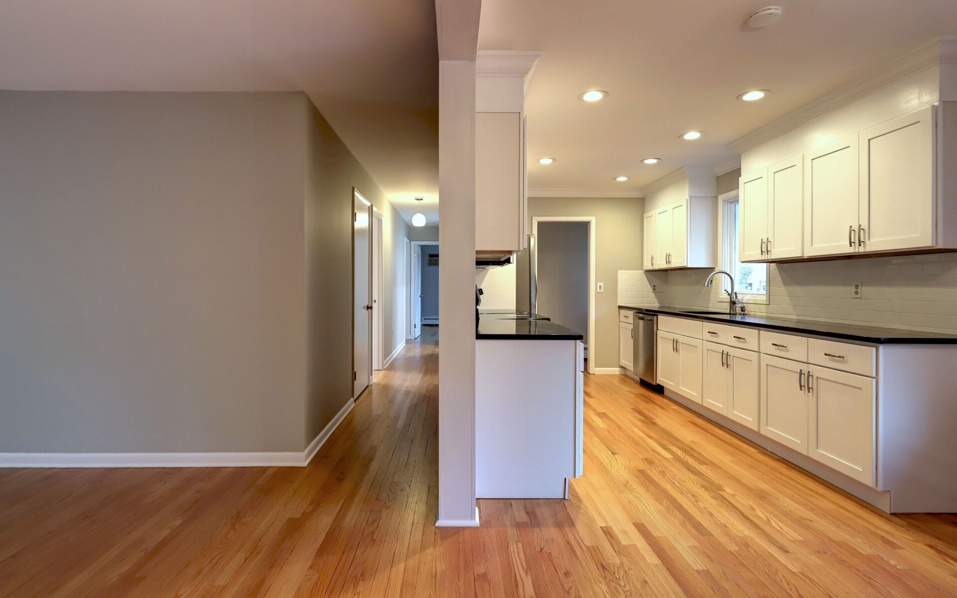 Kitchen with white cabinets, dark countertops, and wood floor. Hallway with gray wall and wooden floor.