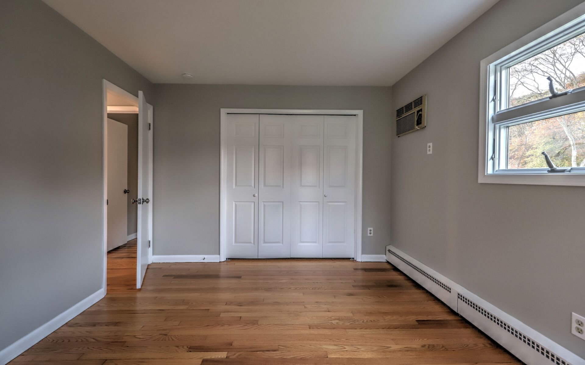 Empty bedroom with hardwood floors, gray walls, white closet, and window.