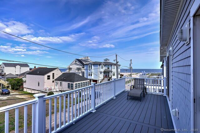 Balcony overlooking a seaside town, blue sky, grey deck with a chair, white railing, and buildings.