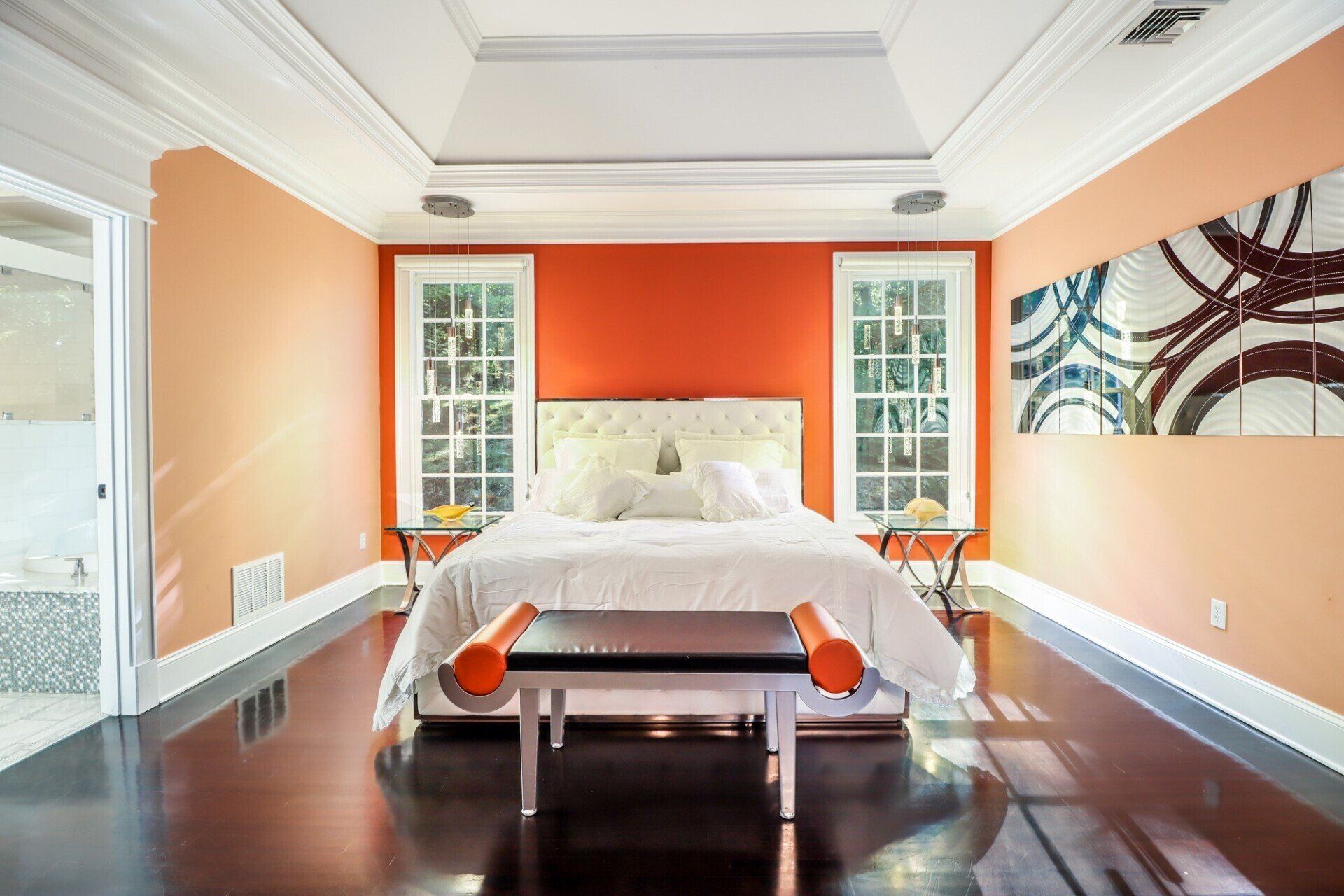 Bedroom with dark wood floors, orange walls, and a white bed. Two windows flank the headboard.