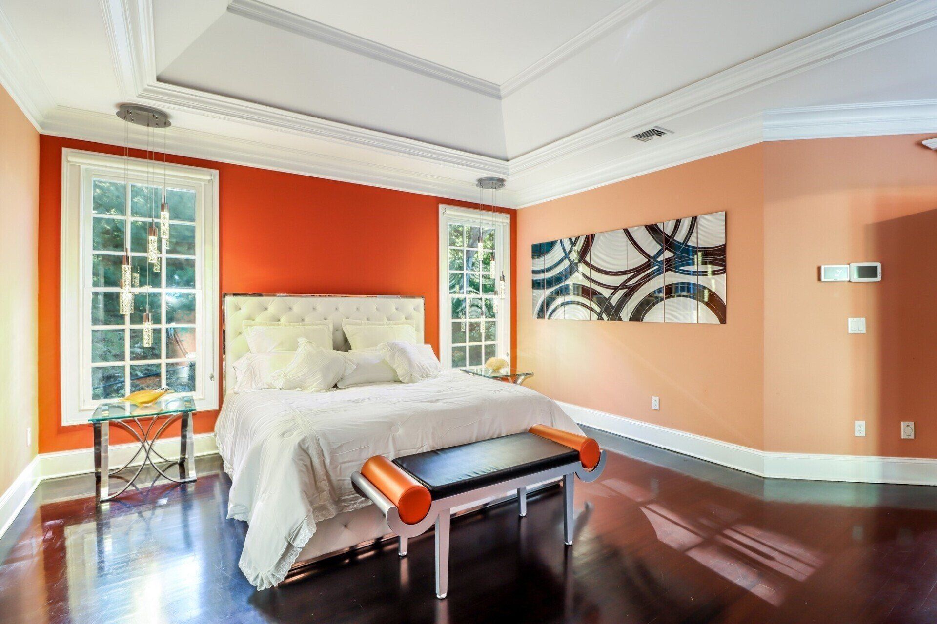 Bedroom with an orange accent wall, bed, windows, artwork, and dark wood floor.