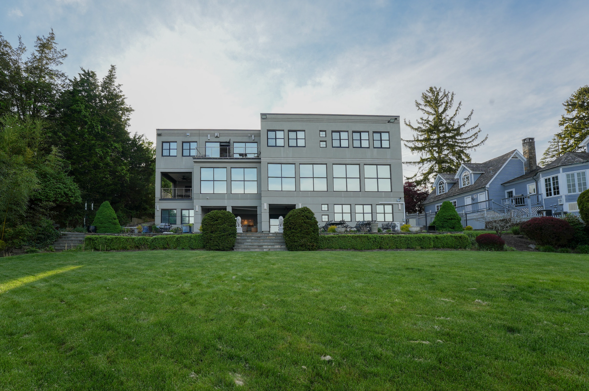 Large gray house with many windows on a green lawn, near trees and water, under a cloudy sky.