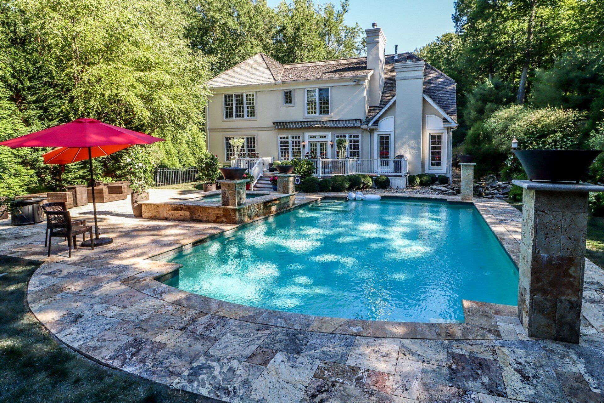 Swimming pool in backyard of a two-story house, with red umbrella, trees, and stone patio.