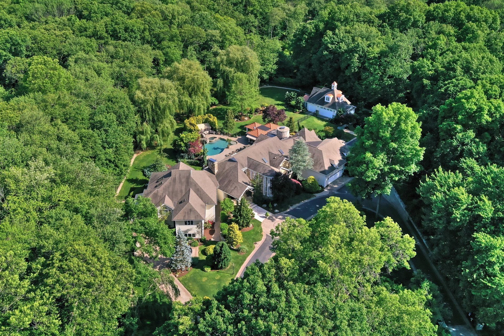 Aerial view of a large house surrounded by lush green trees, with a pool visible in the backyard.