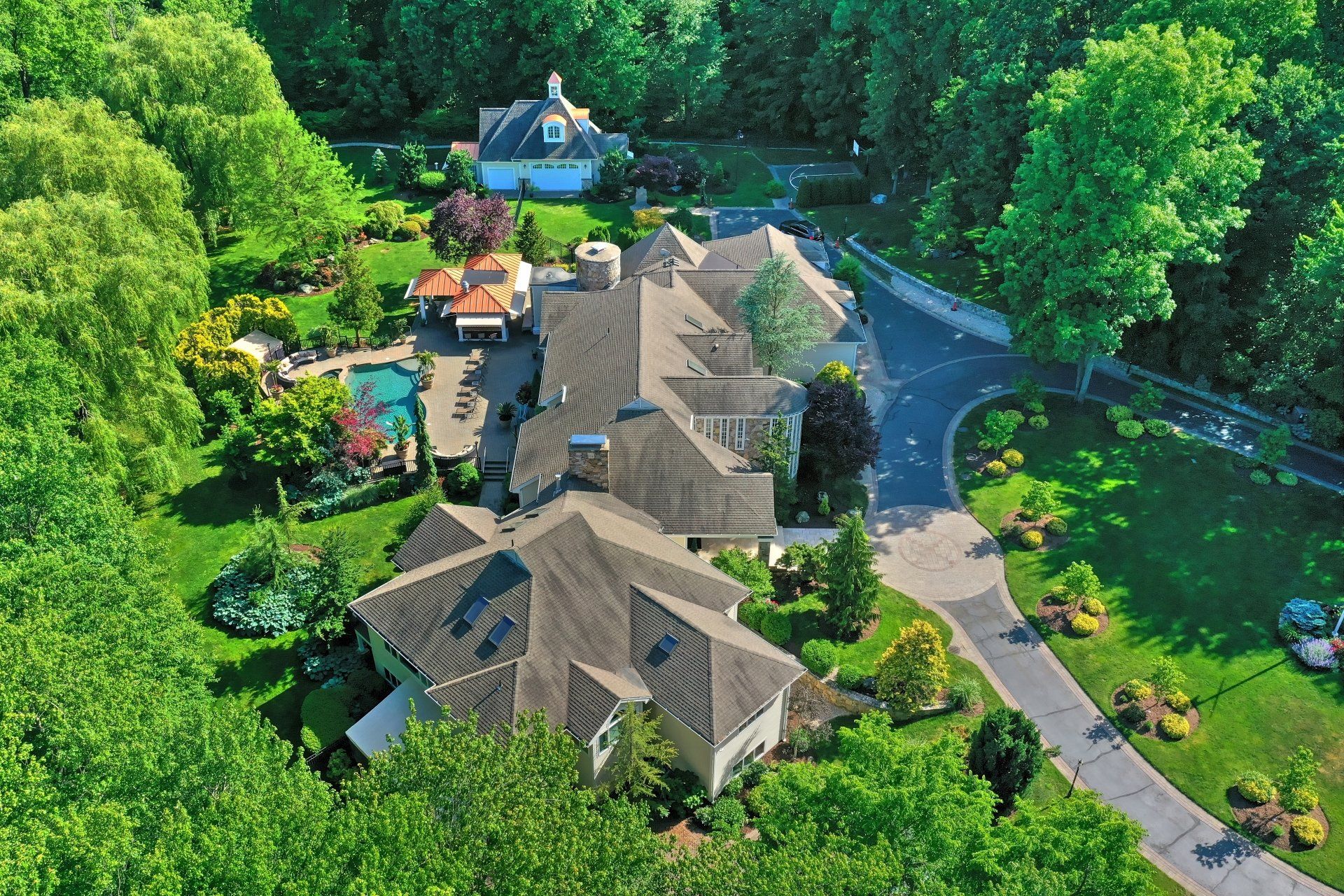 Aerial view of a large house with a pool and lush greenery surrounding it. A long driveway leads to the house.