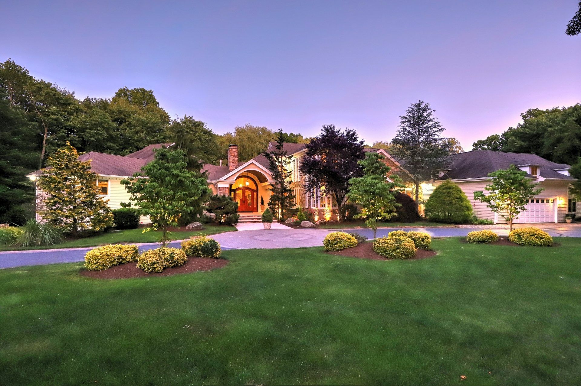 Sprawling, well-lit suburban home with a long driveway and manicured landscaping under a twilight sky.