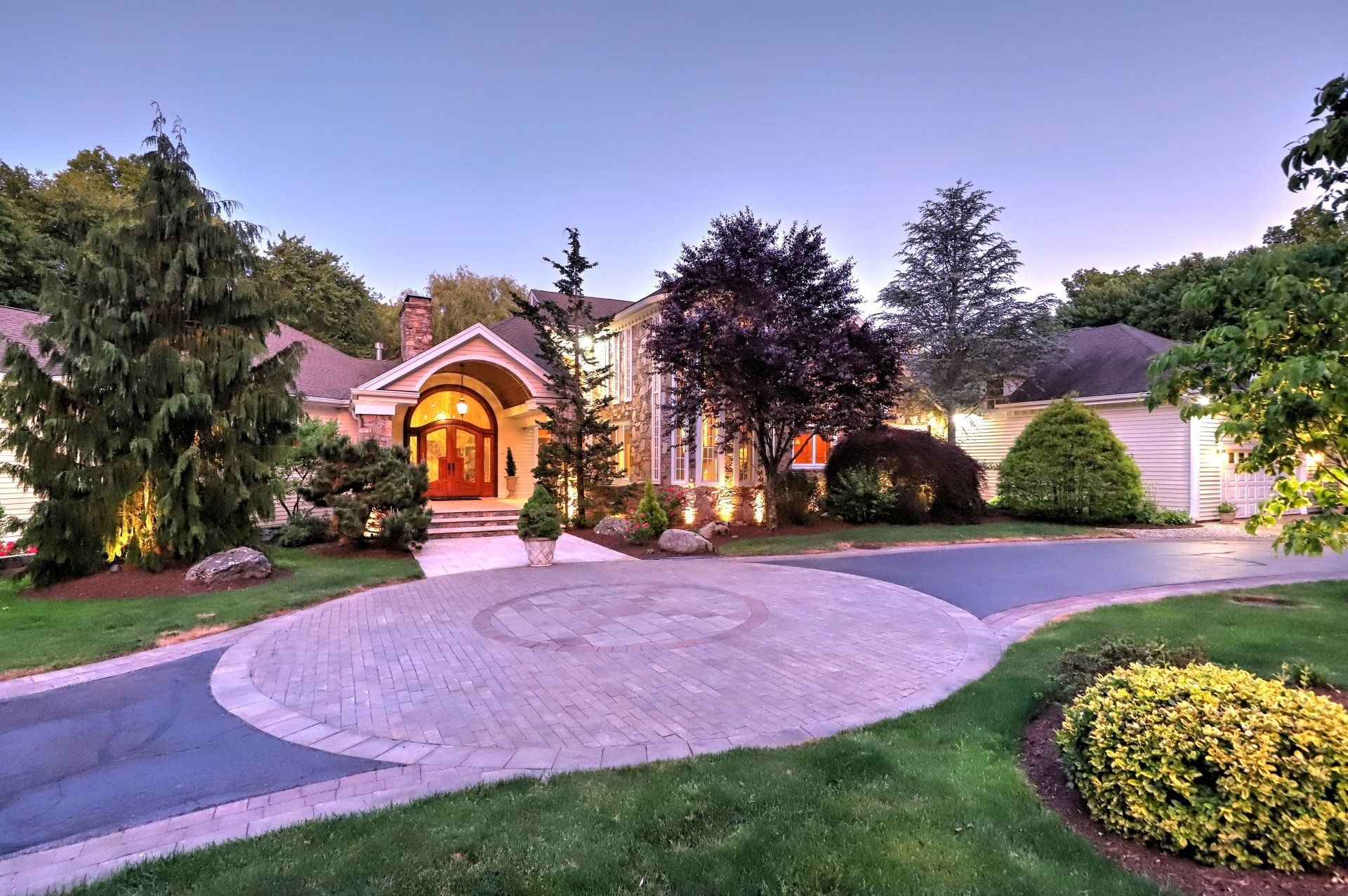 Large house with circular driveway, stone exterior, and manicured landscaping under a twilight sky.