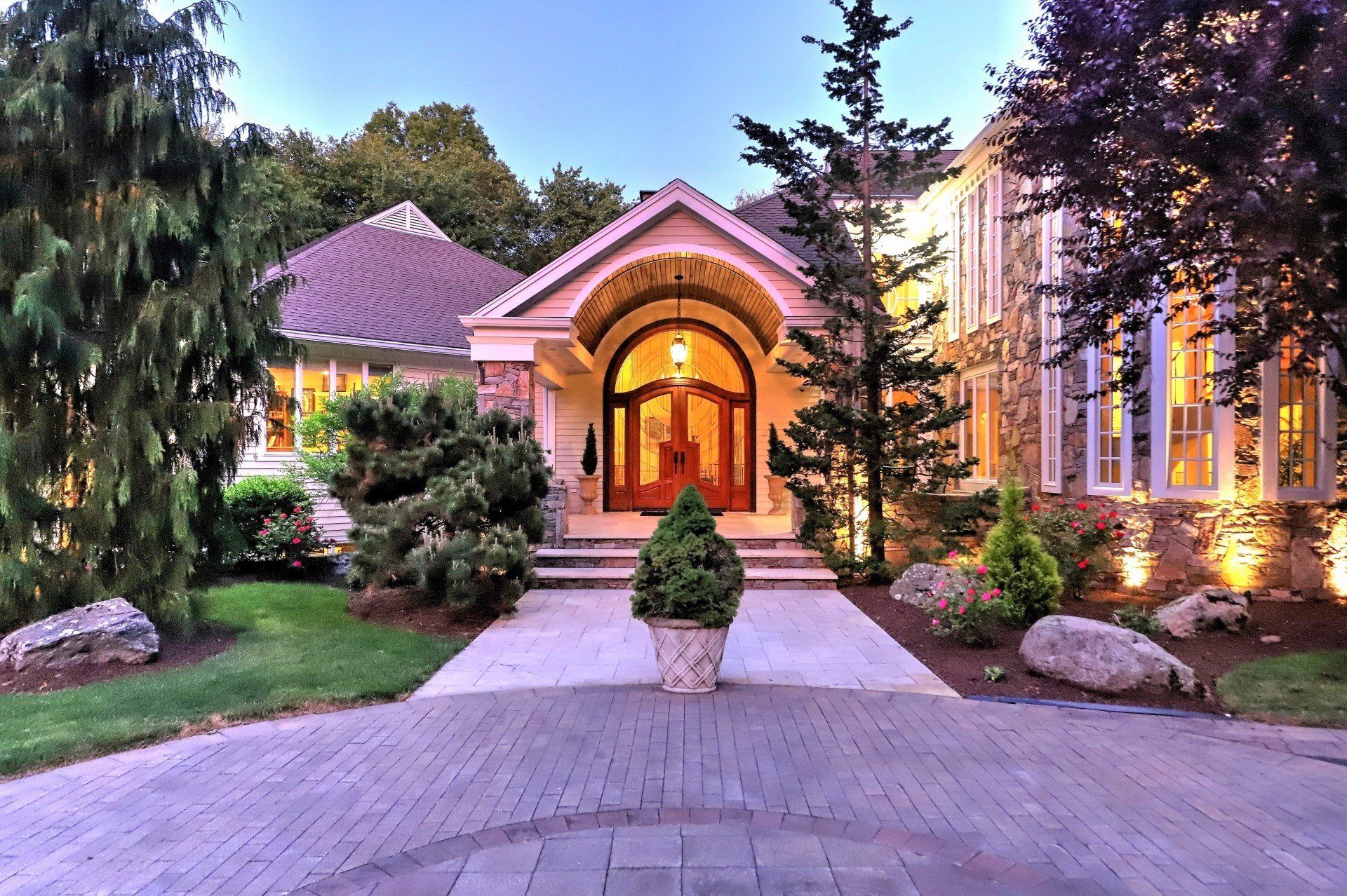 Luxury home with a brick driveway, arched doorway, and lush landscaping.