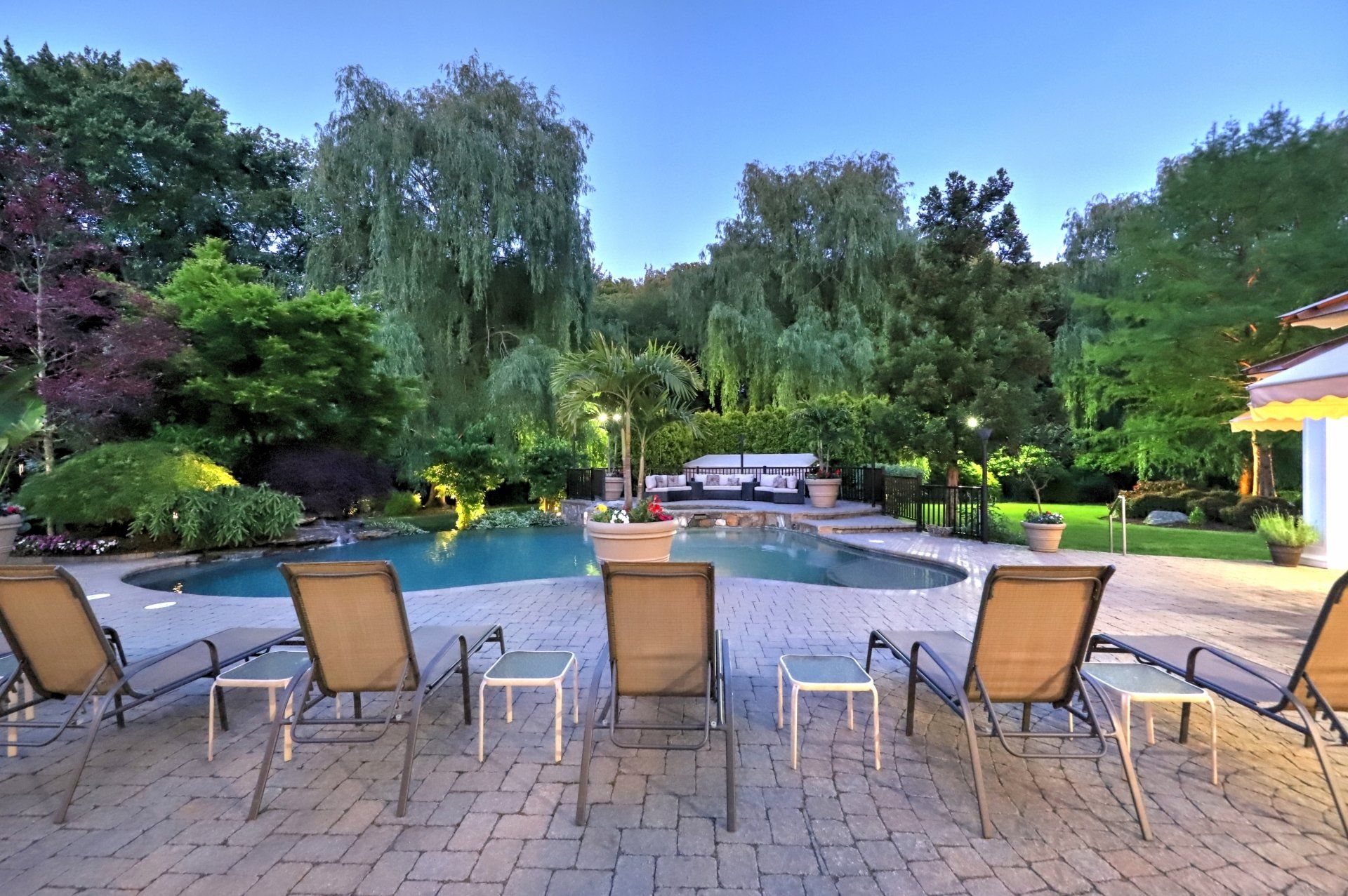 Backyard with pool, lounge chairs, lush greenery, and a trailer under a twilight sky.