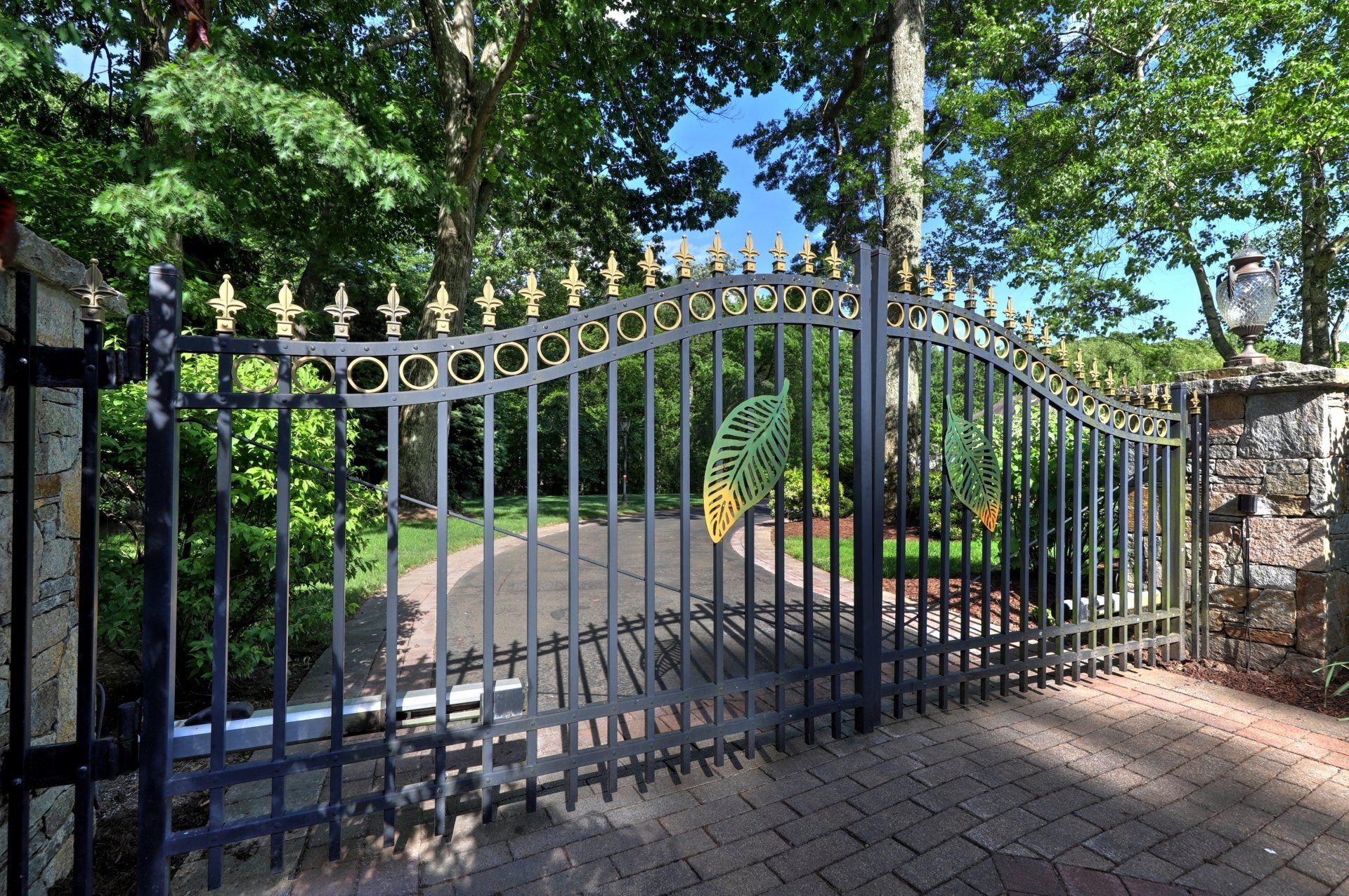 Black iron driveway gate with gold accents, set in a brick and stone structure, with trees in the background.