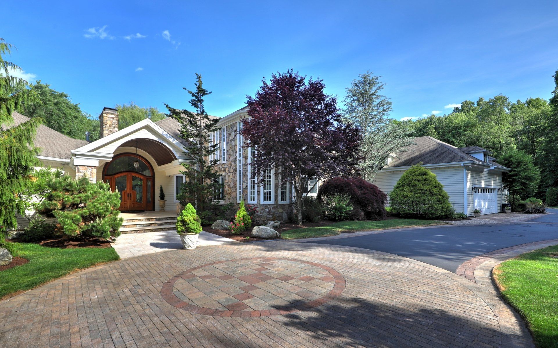 Large stone house with brick driveway, surrounded by trees and green lawn under a blue sky.