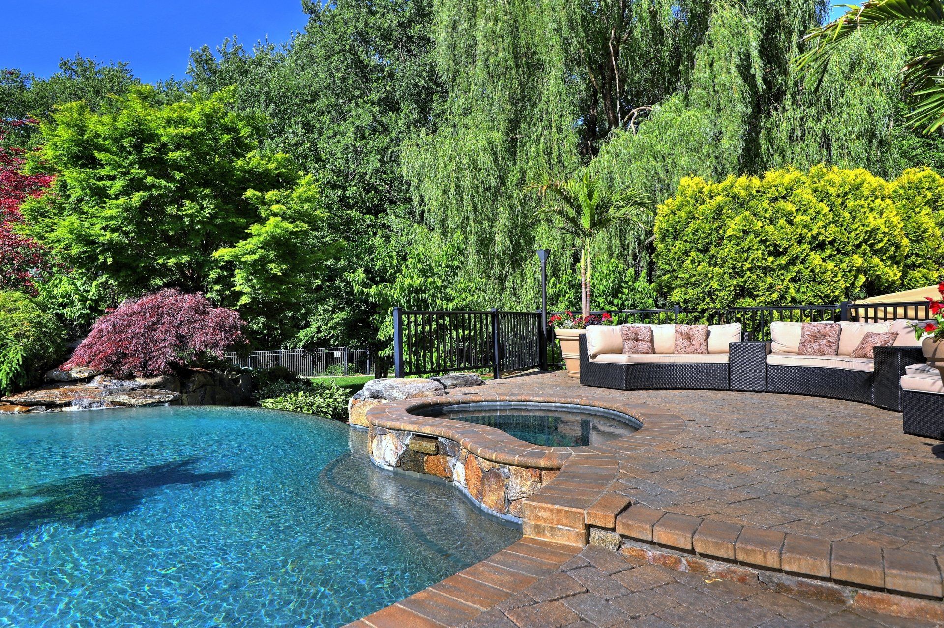 Pool and hot tub with outdoor seating surrounded by lush green trees and foliage.