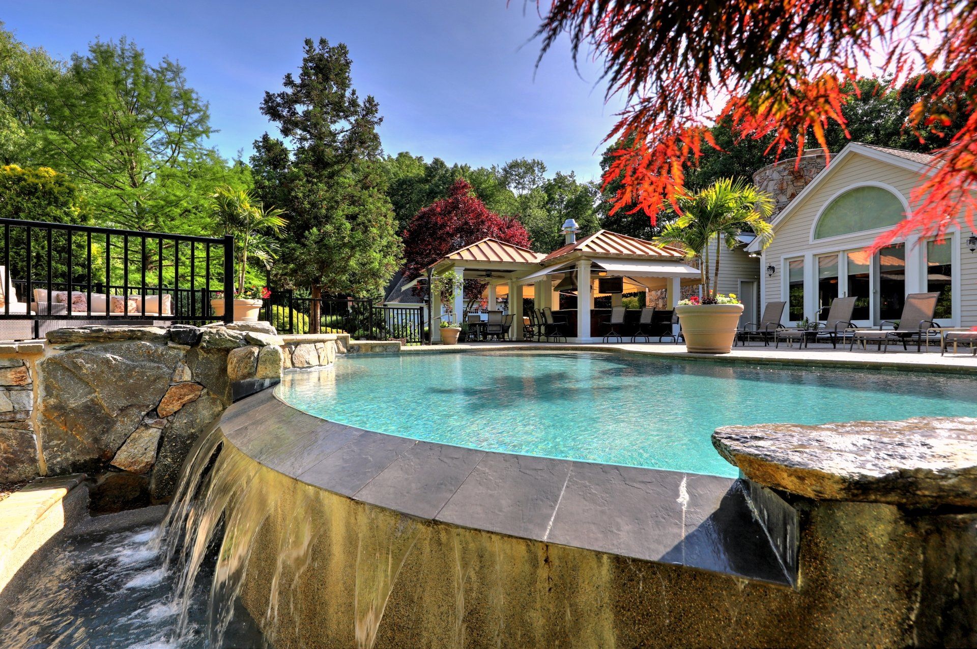 Swimming pool with waterfall, overlooking a gazebo and house, on a sunny day.