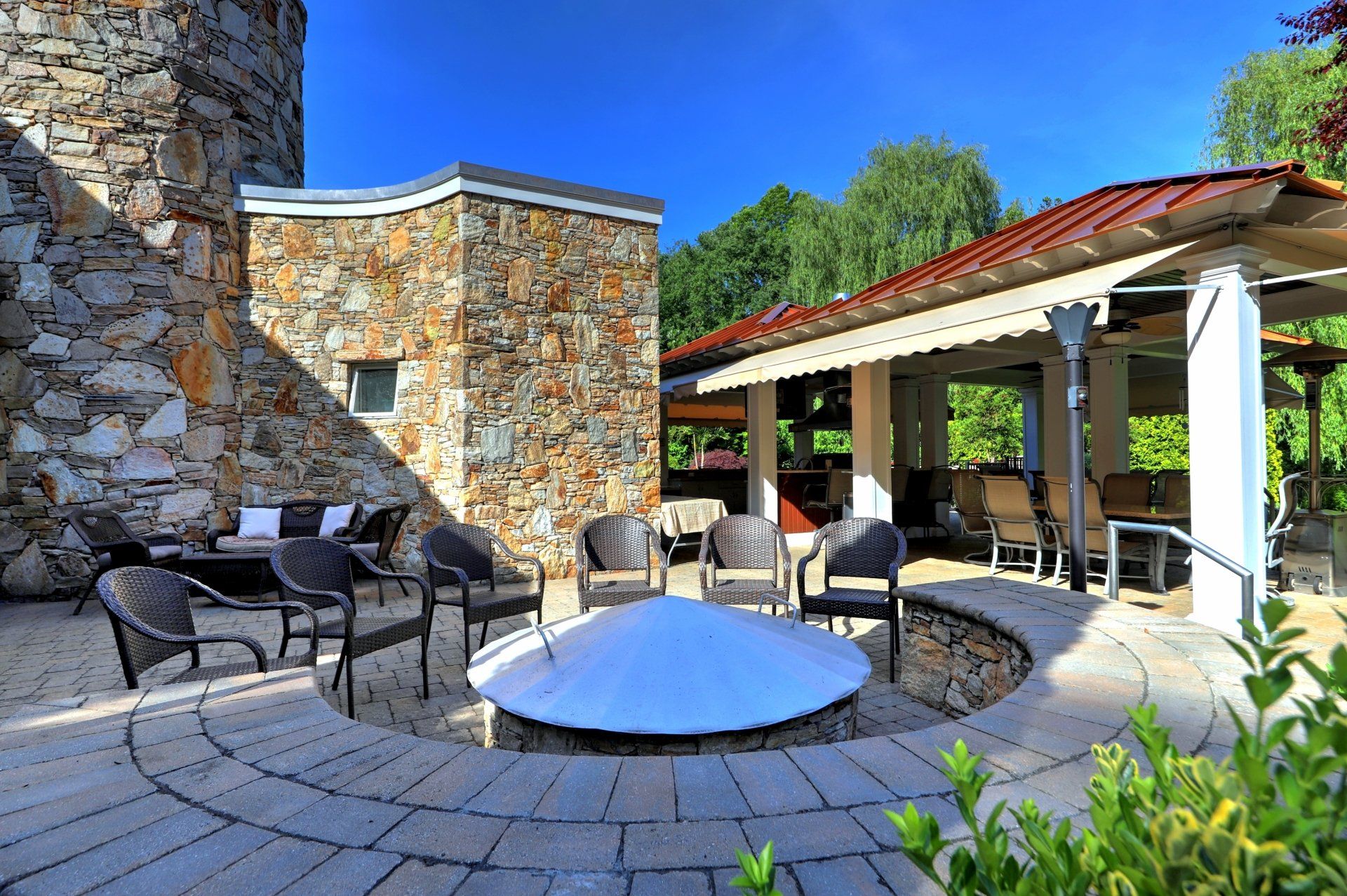 Stone patio with fire pit, seating, and covered patio with red roof under a blue sky.