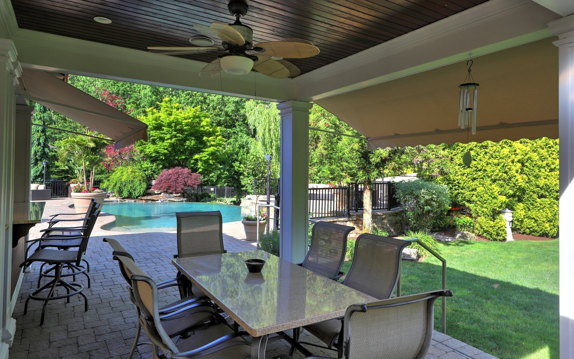 Covered patio with dining table, chairs, and view of pool and lush greenery.