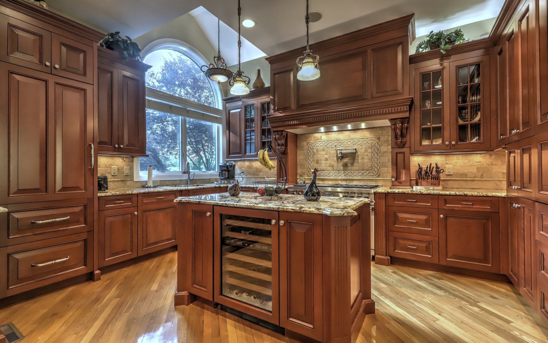 Kitchen with wooden cabinets, island with wine fridge, granite countertops, and large arched window.
