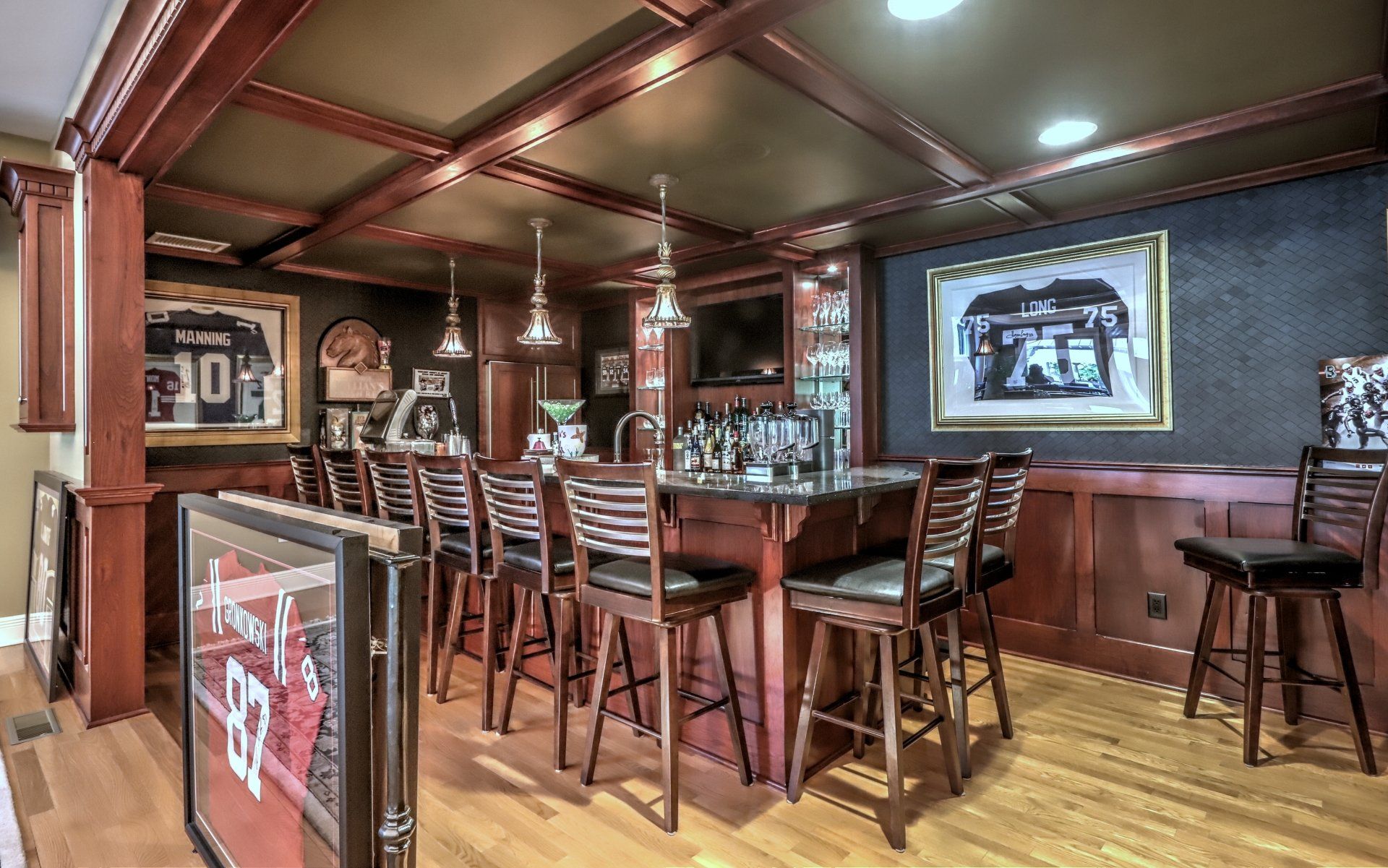 A home bar with dark wood accents, bar stools, sports memorabilia, and overhead lighting.