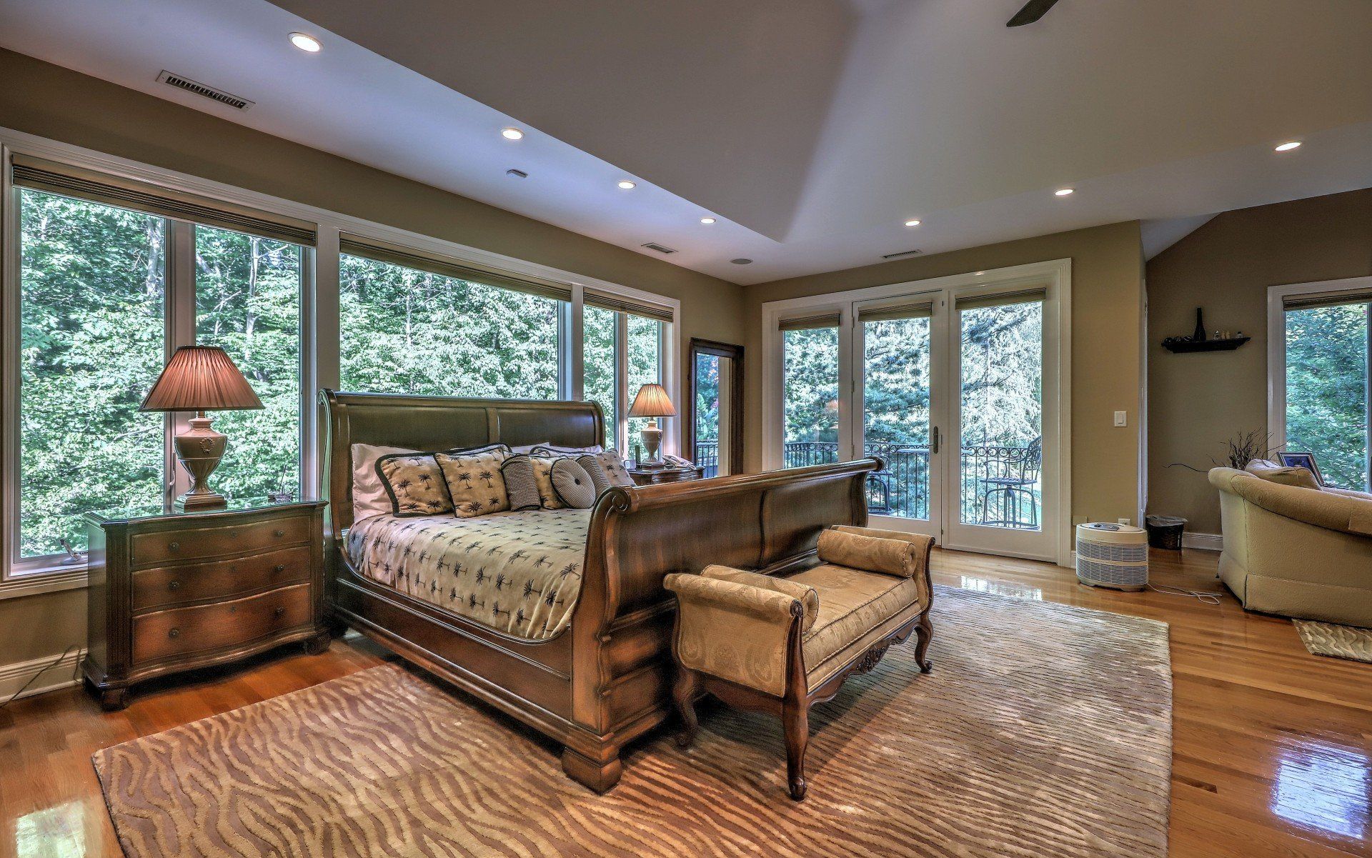 Bedroom with large windows overlooking trees, a bed, seating, and hardwood floors.