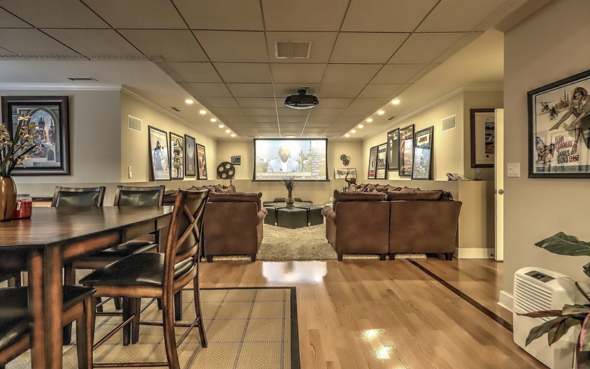 Home theater with projector screen, brown sofas, wooden floor, and framed art on the walls.