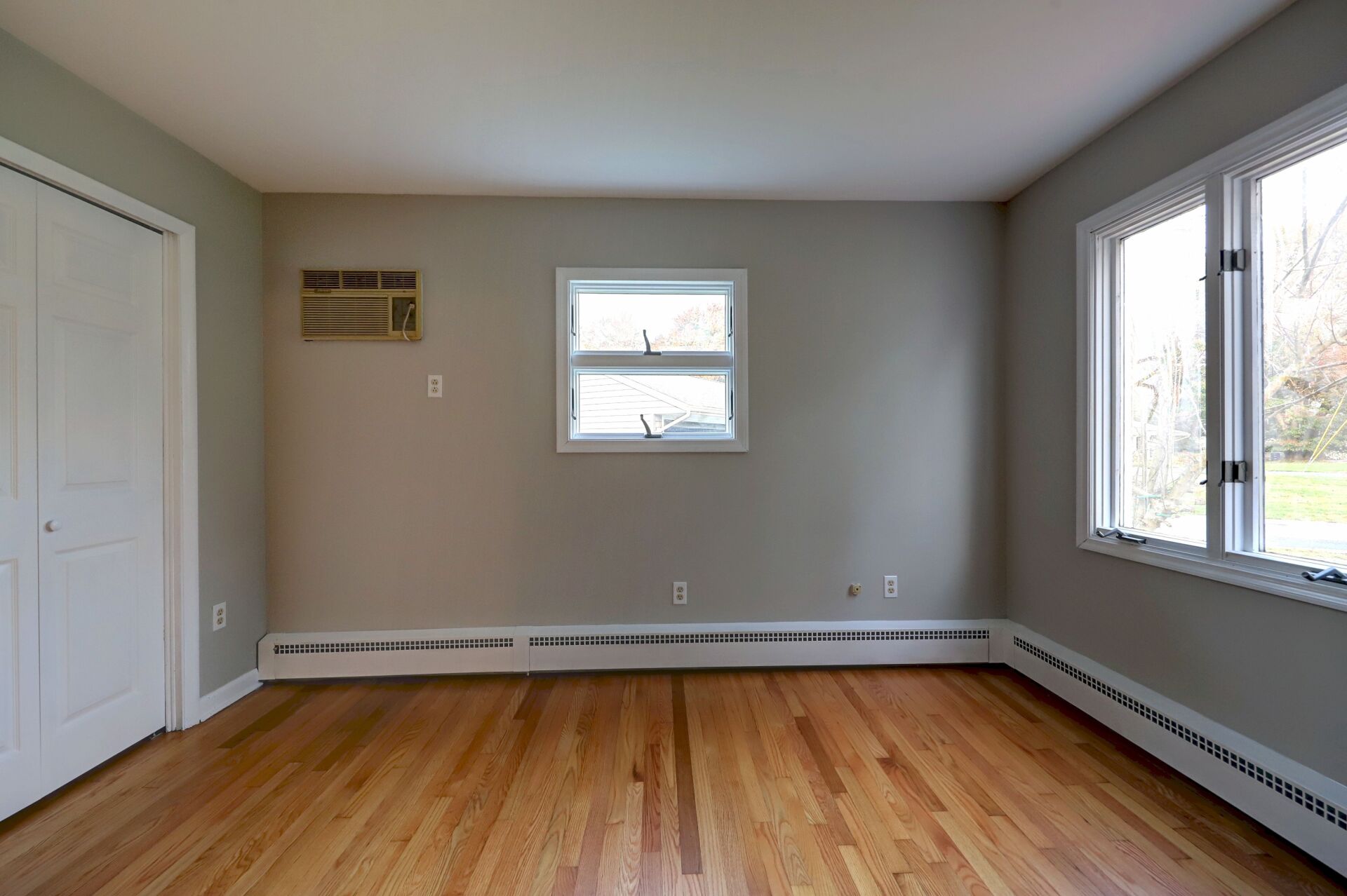 Empty room with hardwood floors, two windows, a closet, and an air conditioning unit on the wall.