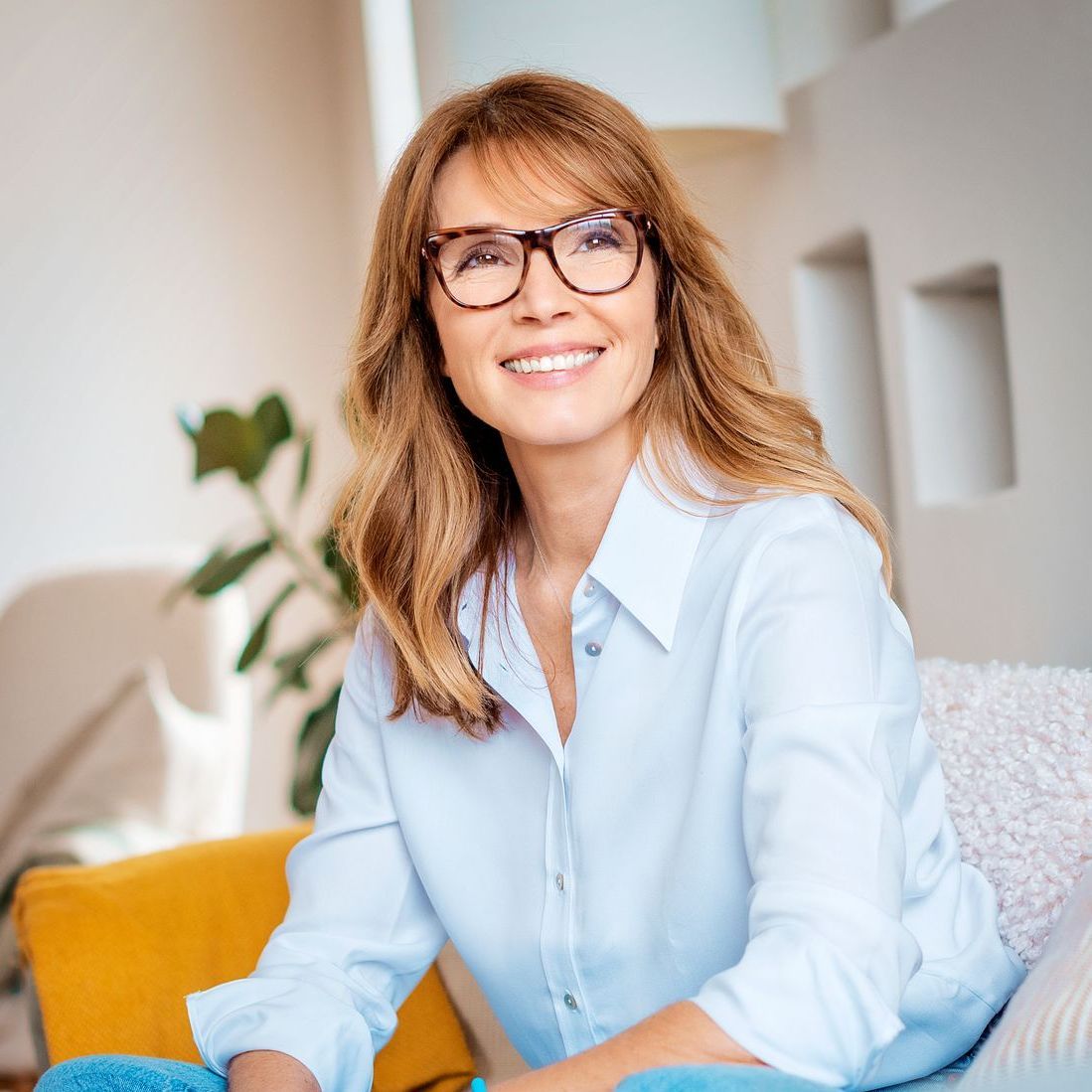 A woman wearing glasses is sitting on a couch and smiling.