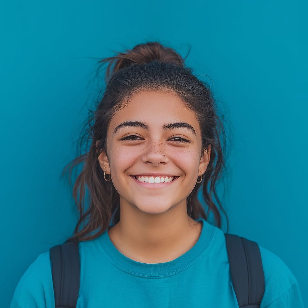 Young woman with dark hair in a bun, smiling broadly in front of a blue background, wearing a blue shirt and backpack.