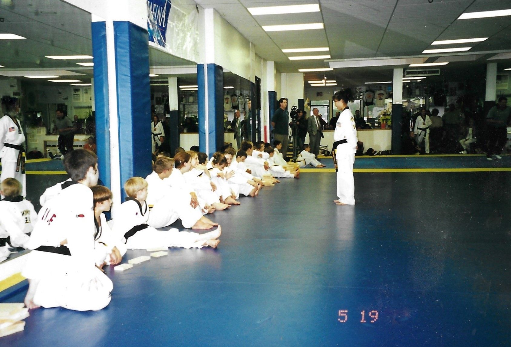 A group of kids are sitting on the floor in a gym with the date 5 19. Mancino Academy Martial Arts