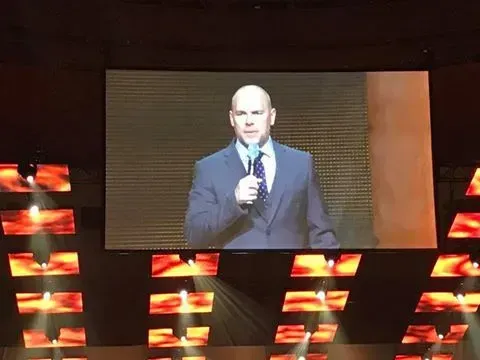 A man in a suit and tie is giving a speech on a large screen