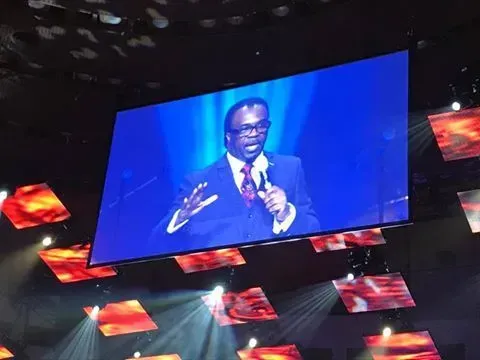 A man in a suit and tie is speaking into a microphone on a large screen