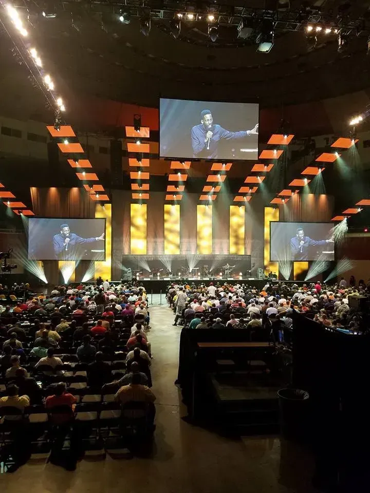 A large auditorium filled with people and a large screen hanging from the ceiling.